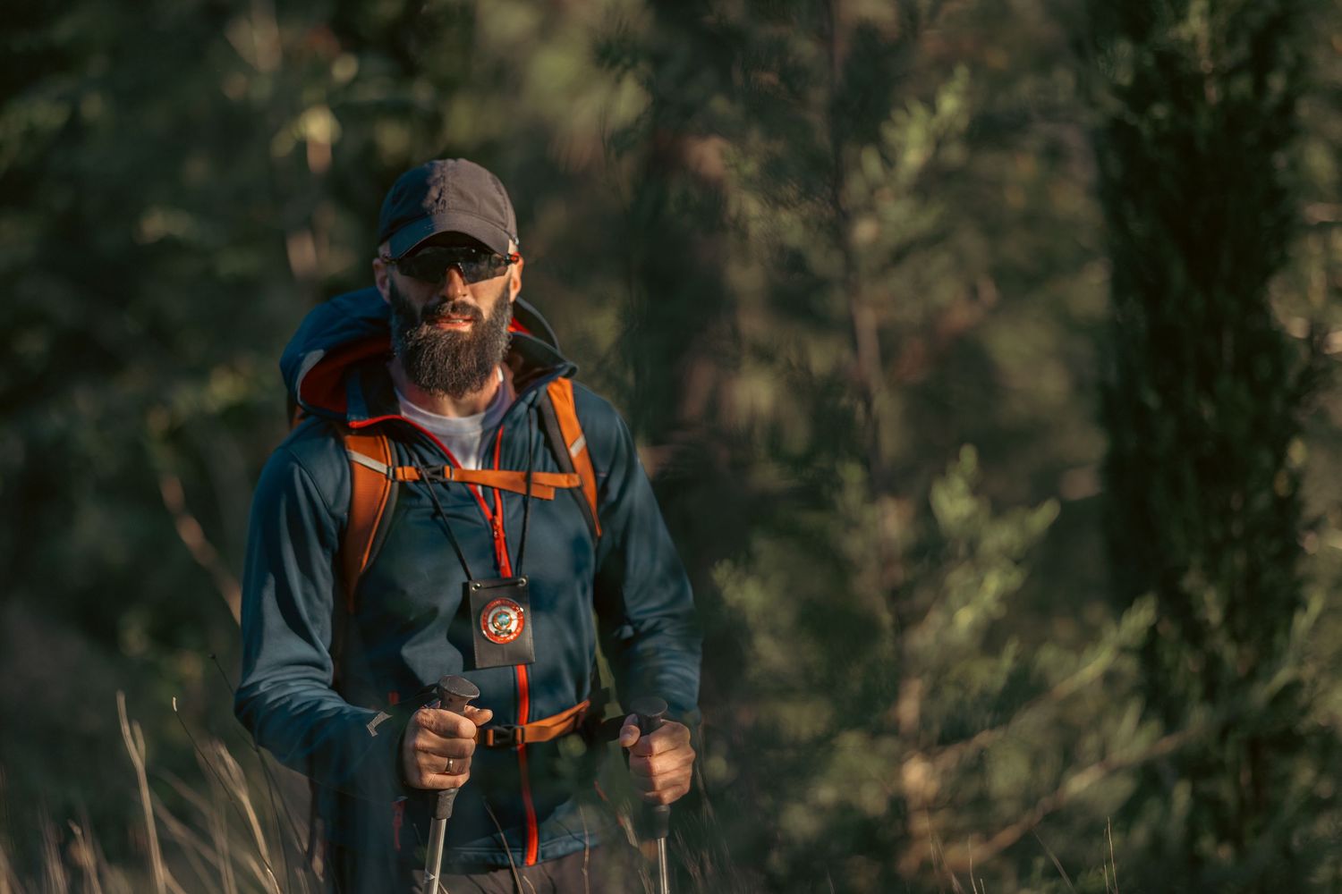 Bearded male hiker with orange backpack and trekking poles walking through a sunlit forest trail.