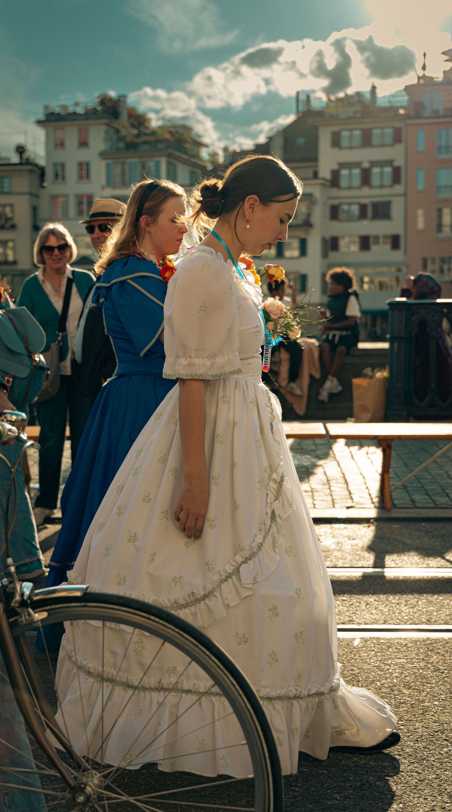 Women in vintage white and blue dresses walk near a bicycle on a sunny European city street.