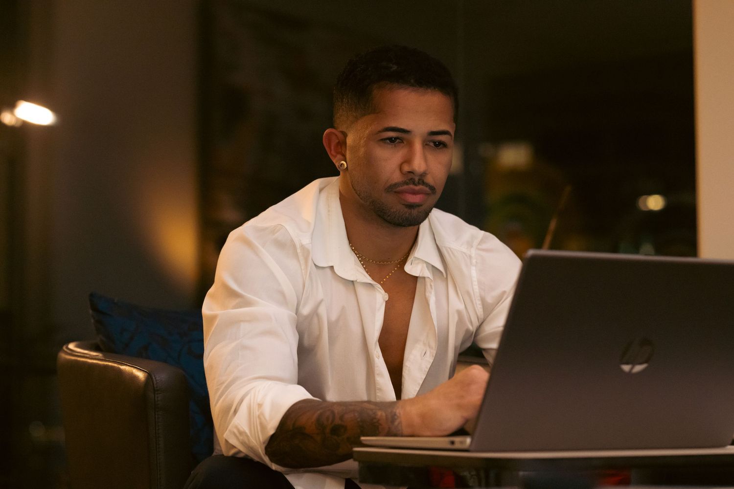 Man in white shirt working on laptop at night, looking focused and serious in dimly lit room.