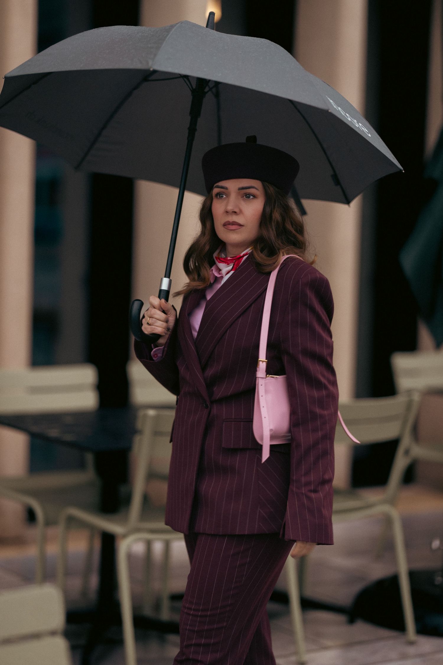Stylish person in burgundy pinstripe suit and beret holding gray umbrella with pink shoulder bag in rainy weather.