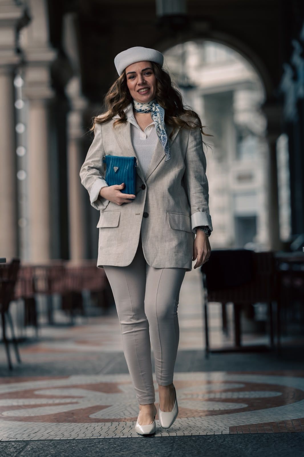 Stylish woman in beige blazer, white beret, and heels holding a blue clutch while walking through an elegant arcade.