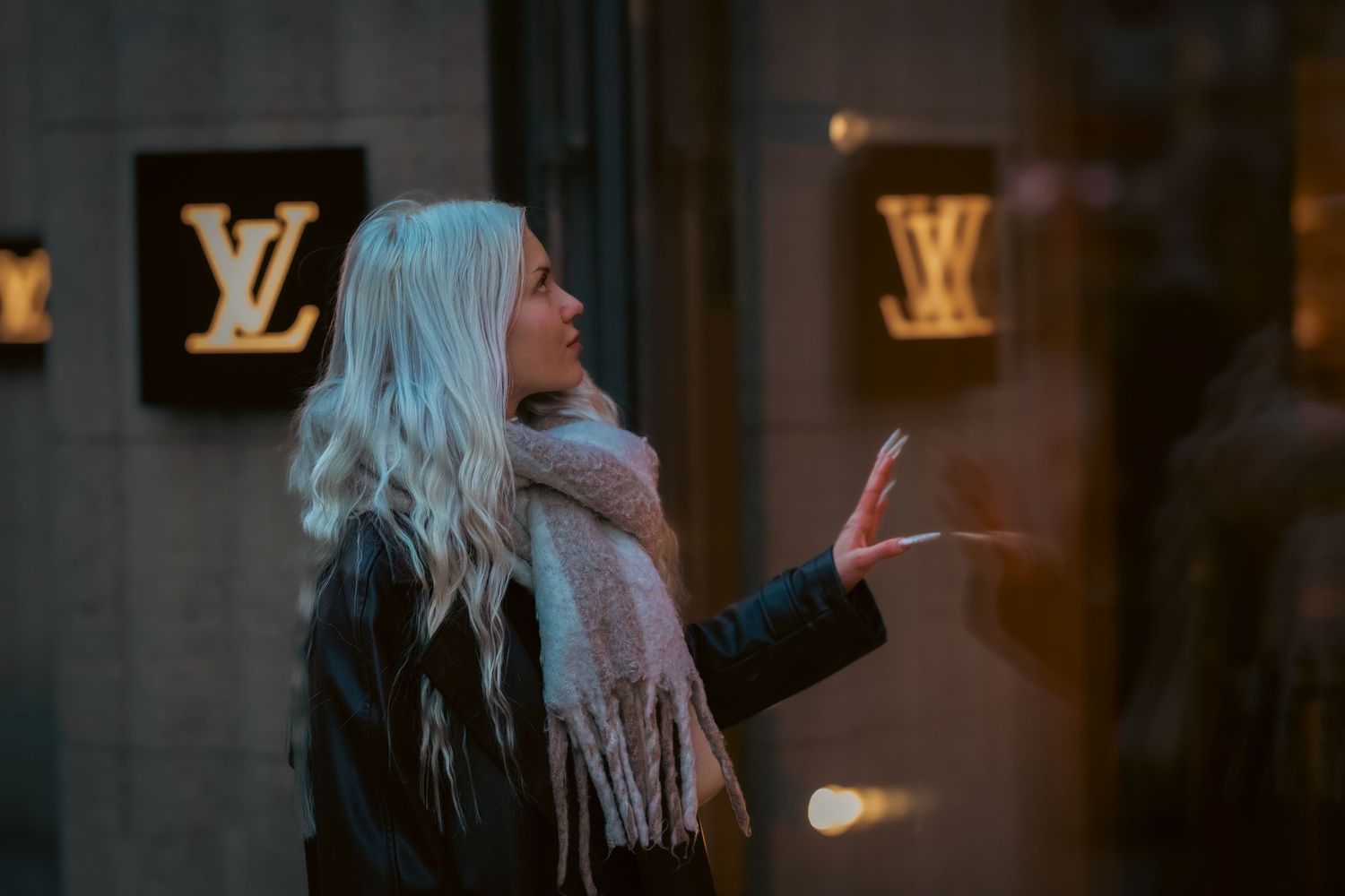 A person with long platinum blonde hair stands in front of illuminated Louis Vuitton store signage at night.
