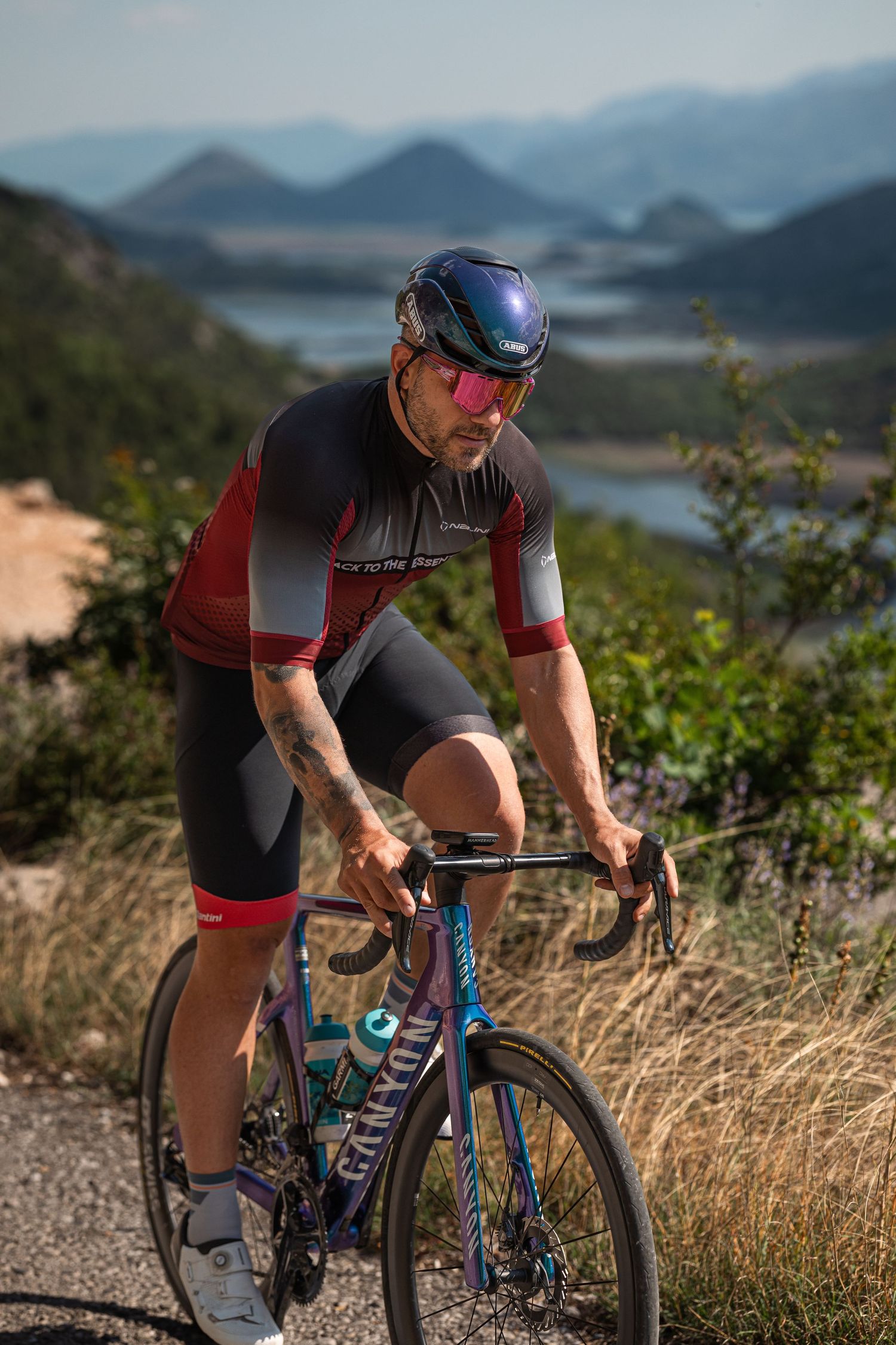 Cyclist in red and black jersey climbs mountain road on colorful bike with scenic lake and hills backdrop.