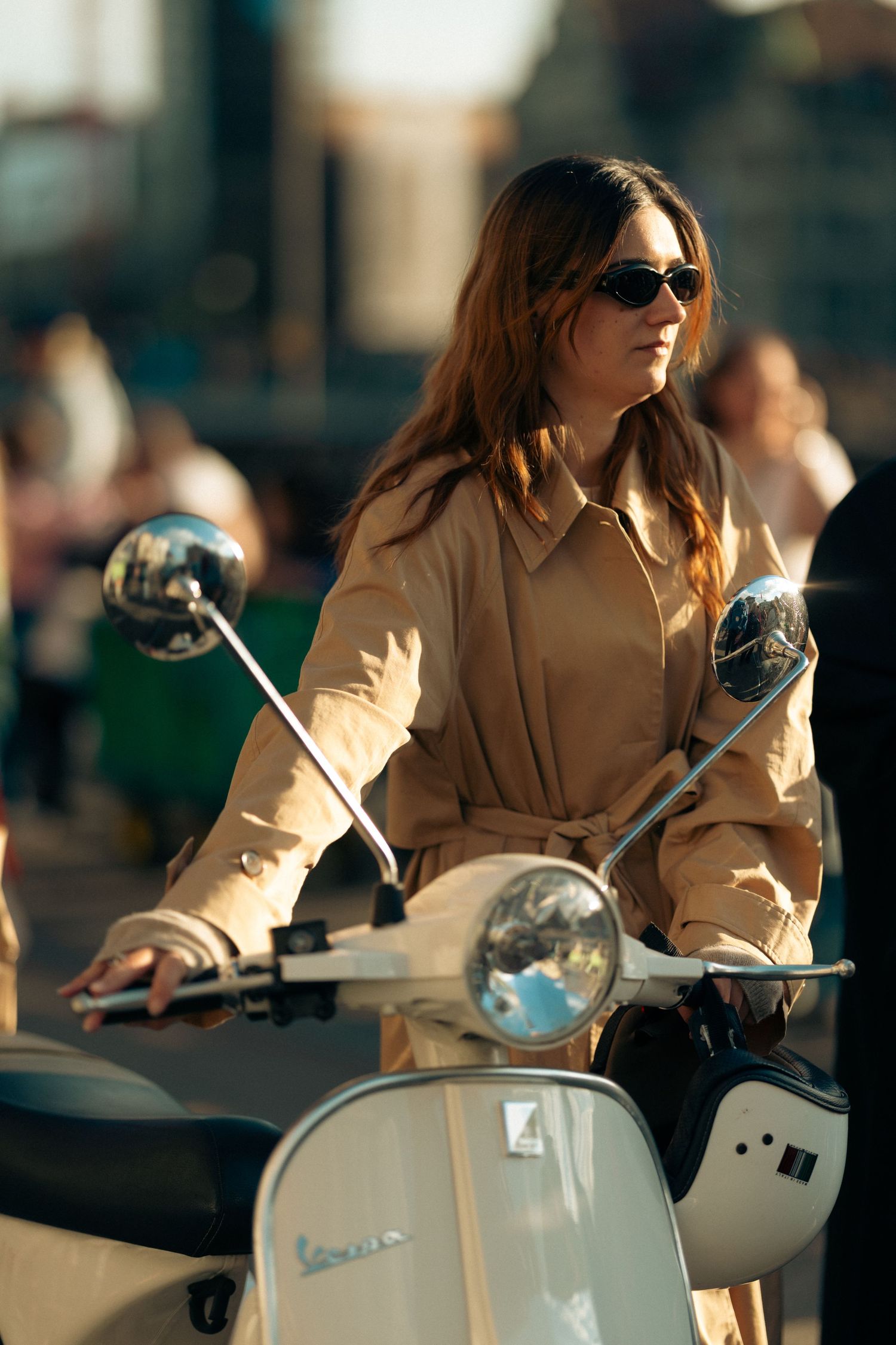 Woman in beige trench coat and sunglasses riding a white Vespa scooter on a sunny urban street.