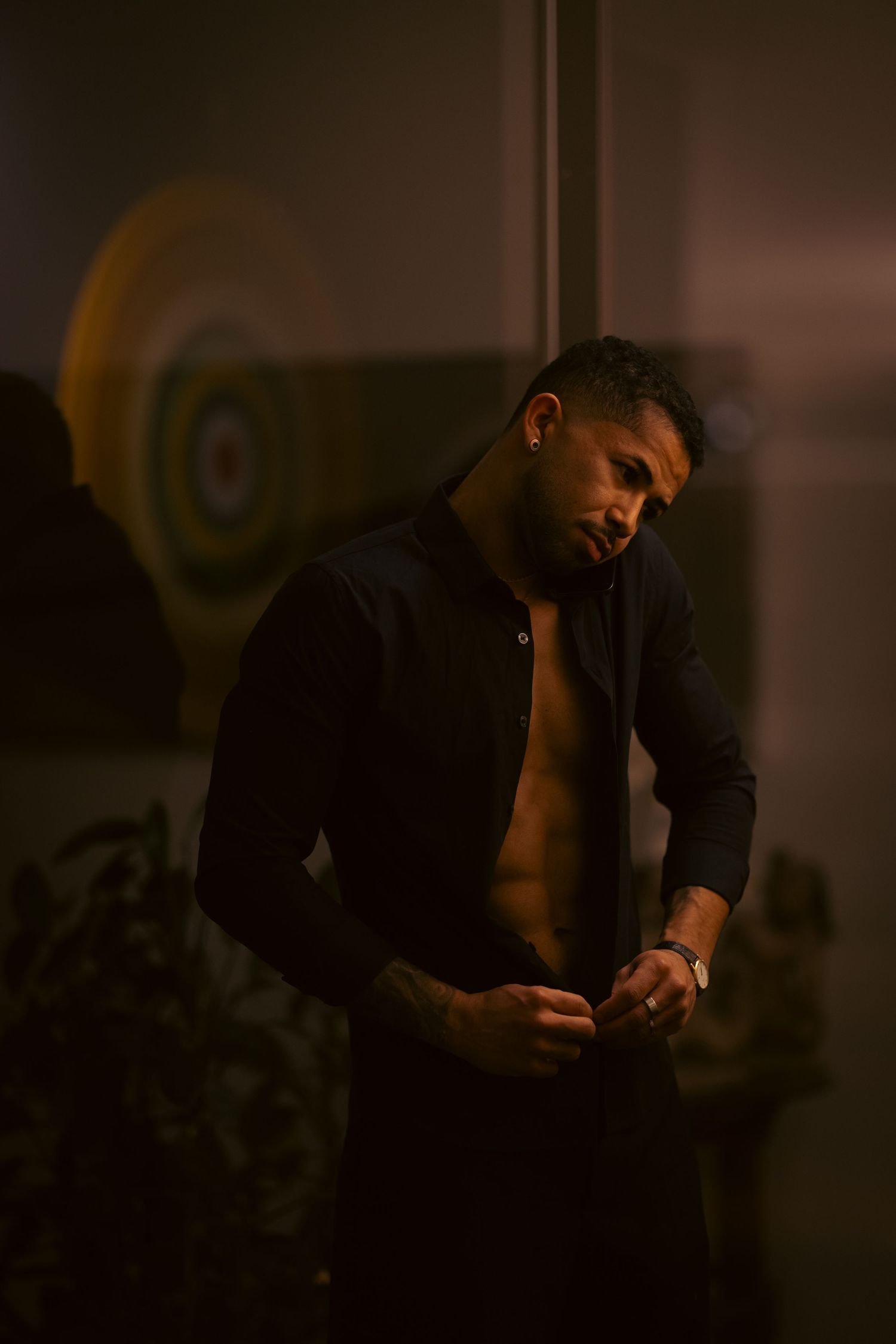 A man in an open black shirt poses in dramatic low lighting, showcasing a moody, stylish aesthetic.