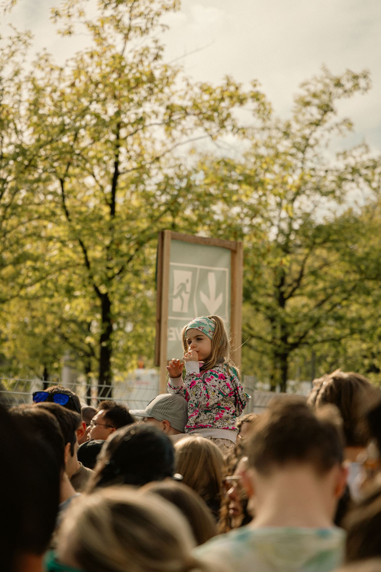 A young girl sits on shoulders above a crowd at an outdoor festival, sipping a drink with trees in the background.
