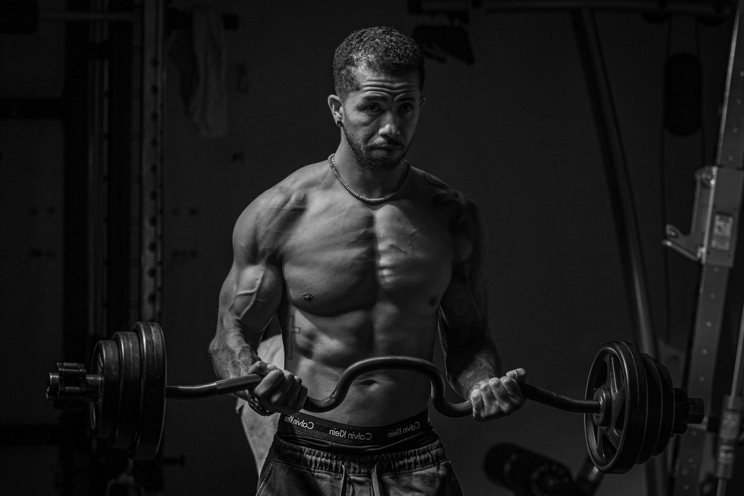 Muscular shirtless man lifting a barbell in a gym, captured in dramatic black and white photography.