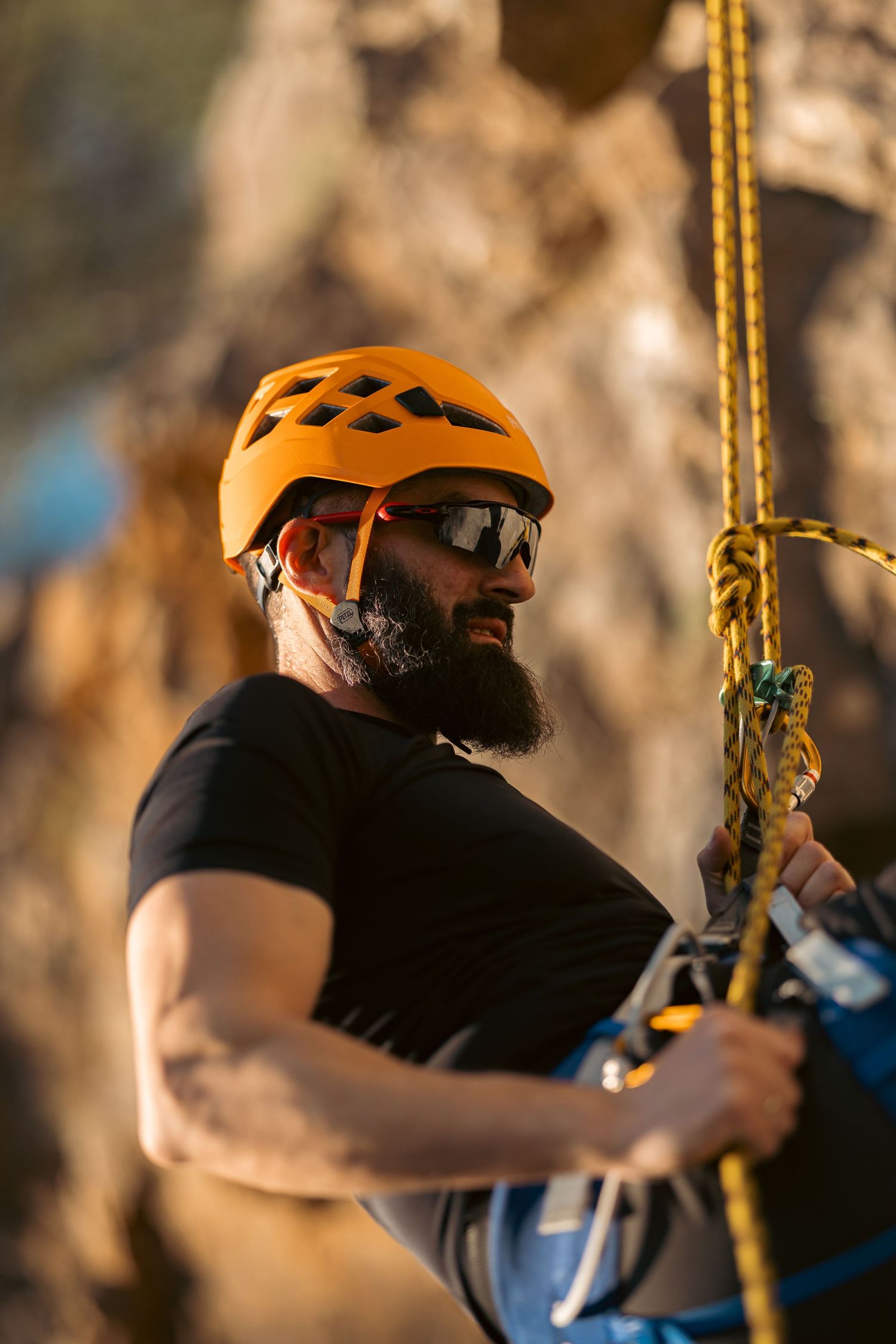 Bearded male rock climber in orange helmet and sunglasses managing rope equipment on a cliff face.