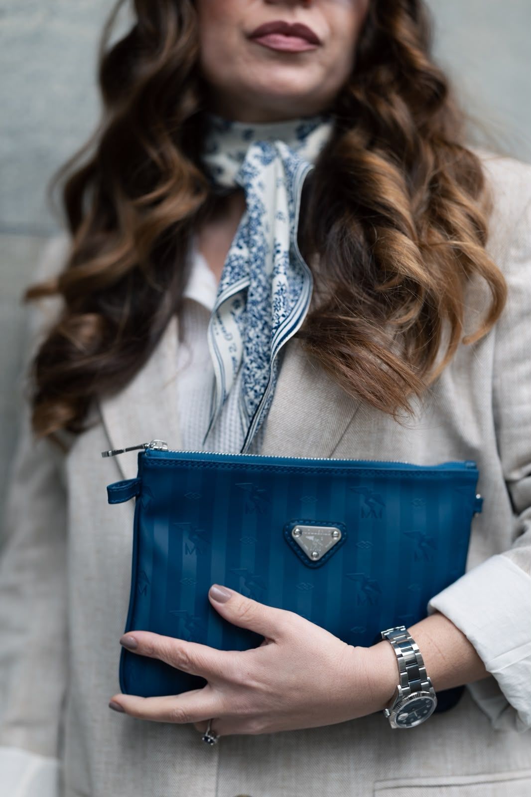 Woman holding a blue Prada clutch bag, wearing a silver watch, scarf, and beige blazer.