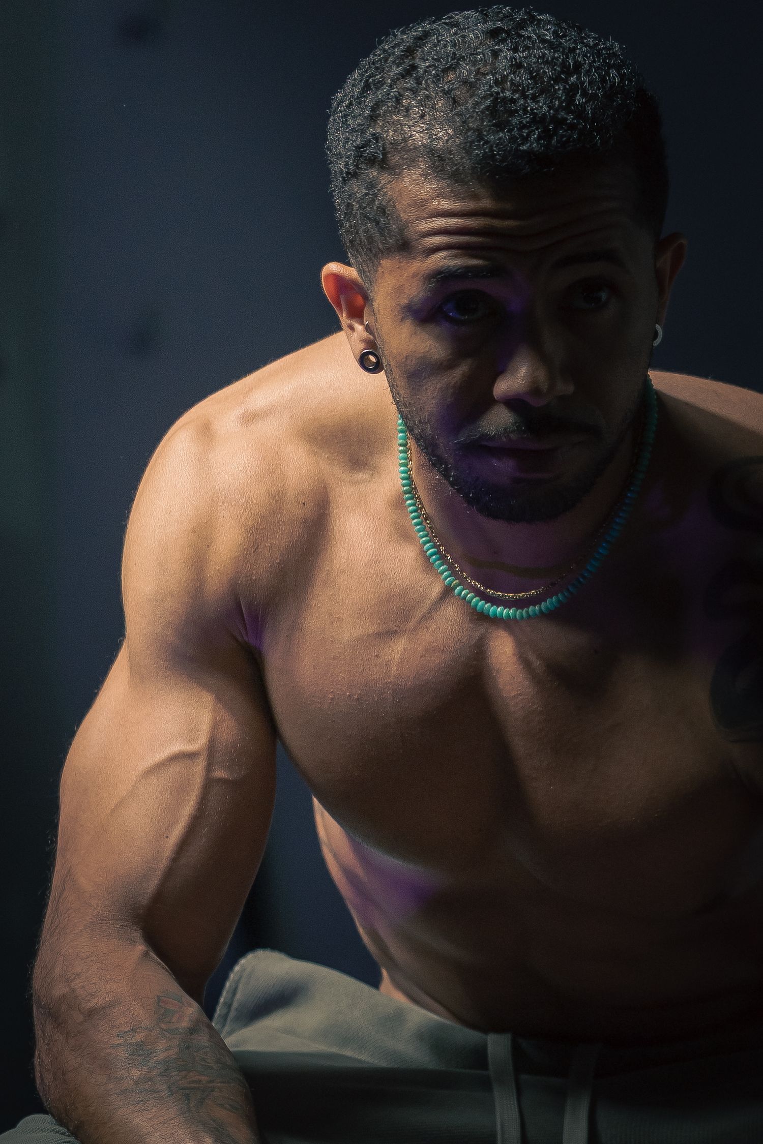 Muscular shirtless man wearing turquoise beaded necklace and ear gauge, posing confidently in dramatic studio lighting.
