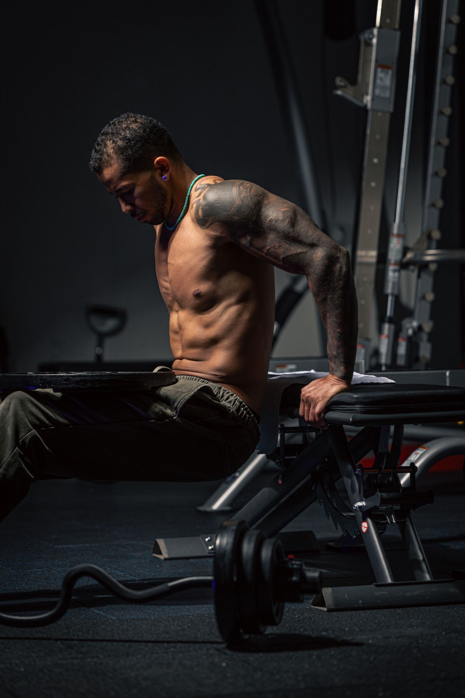 Tattooed shirtless muscular man resting on gym bench during intense workout session in dark fitness center.