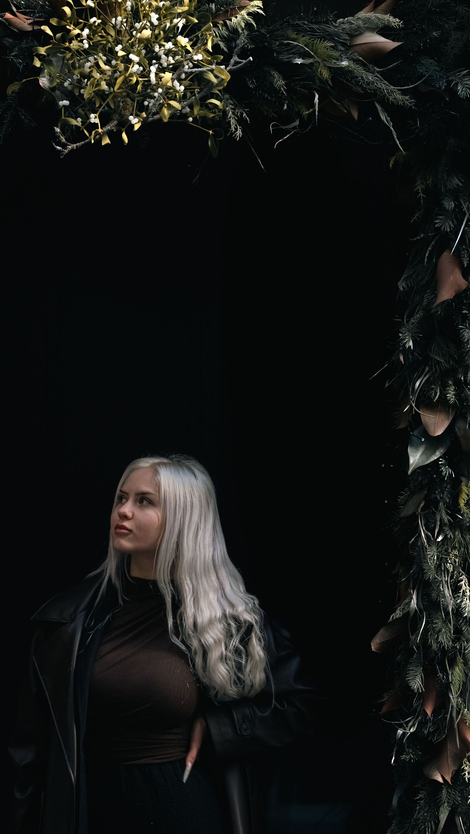 A dark moody portrait with platinum blonde hair framed by green foliage and shadows against a black background.