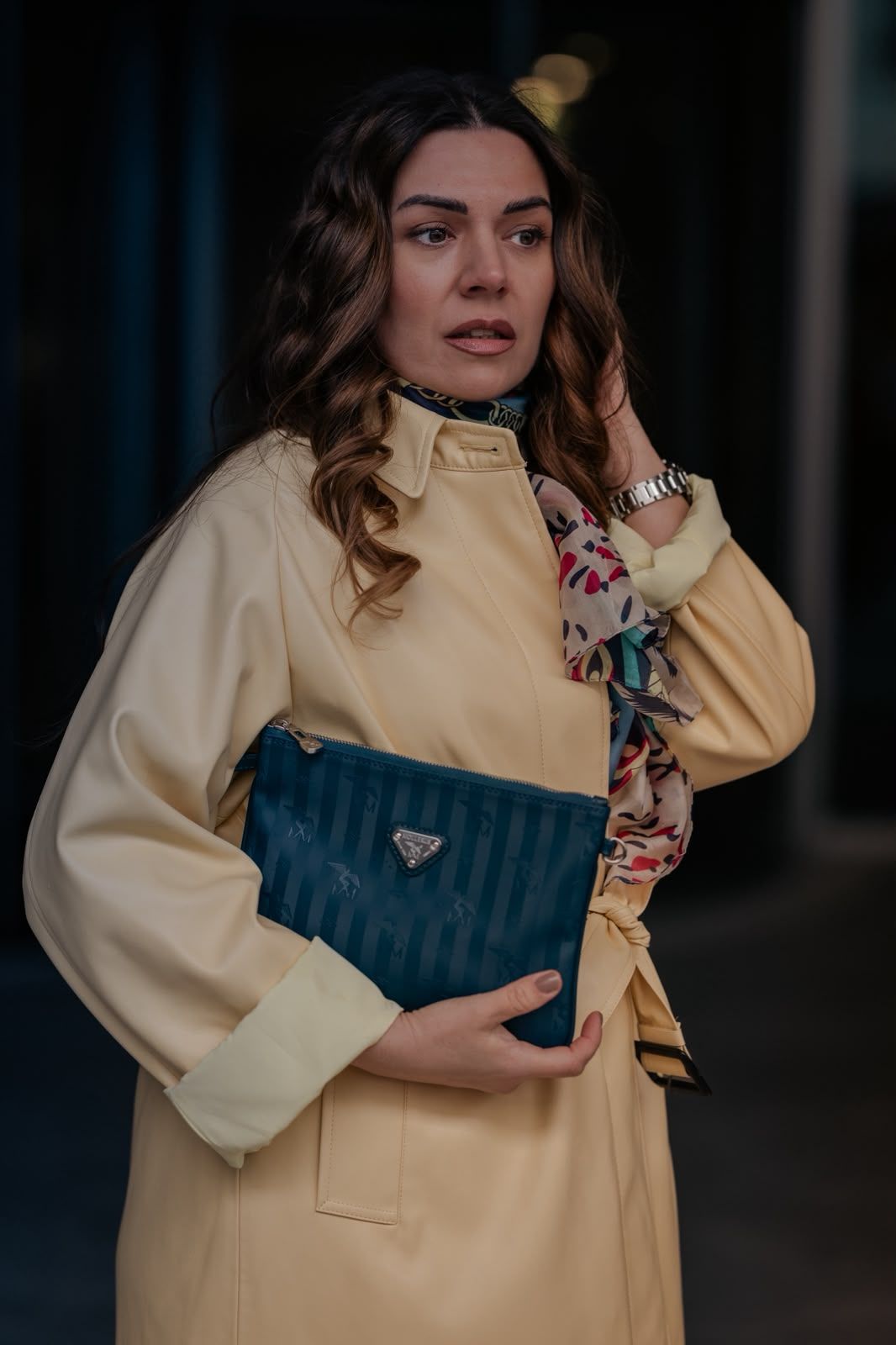 Woman in yellow coat holding a teal Prada clutch bag, wearing a floral scarf and silver watch.