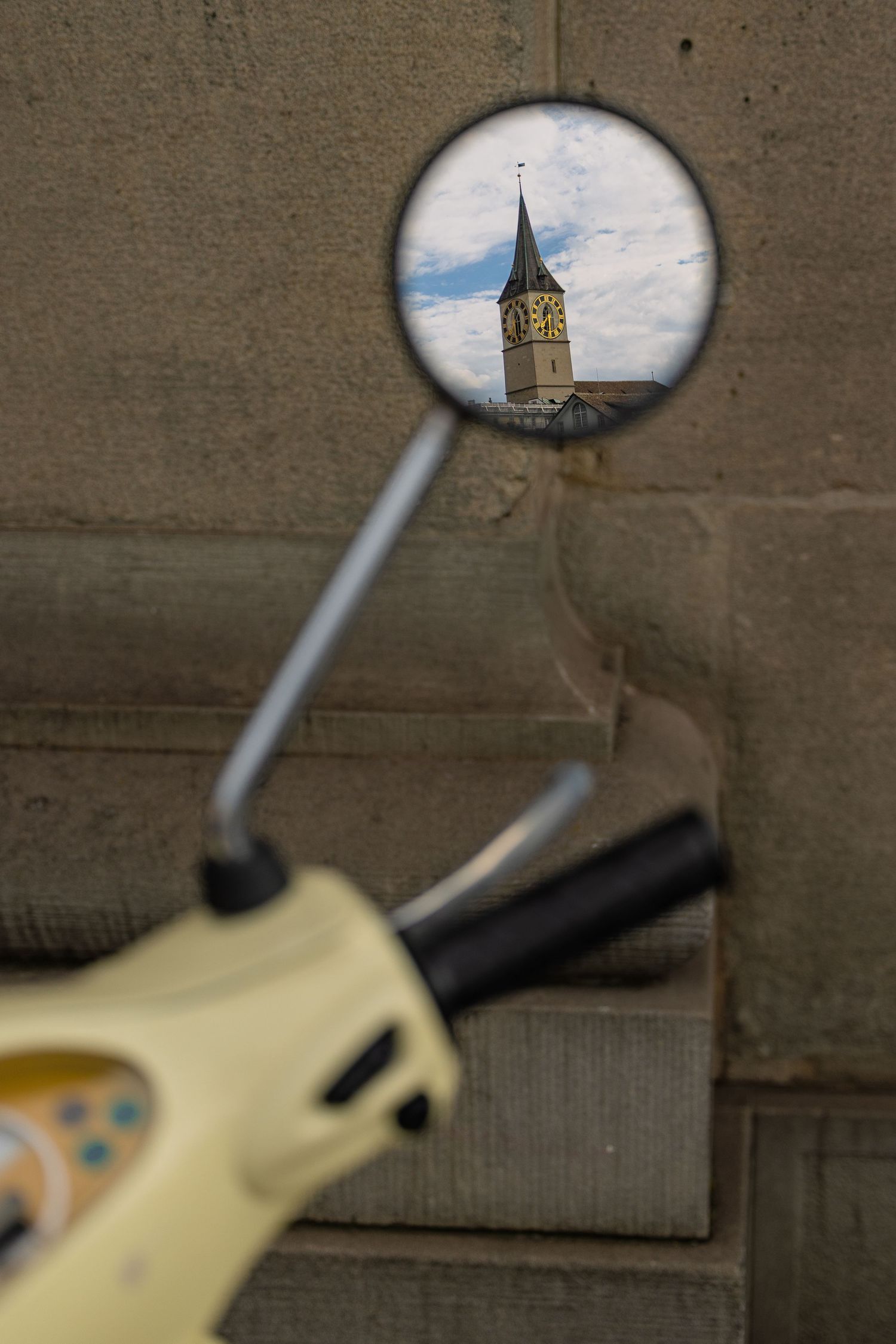 Church tower reflected in a scooter's side mirror against a stone wall backdrop.