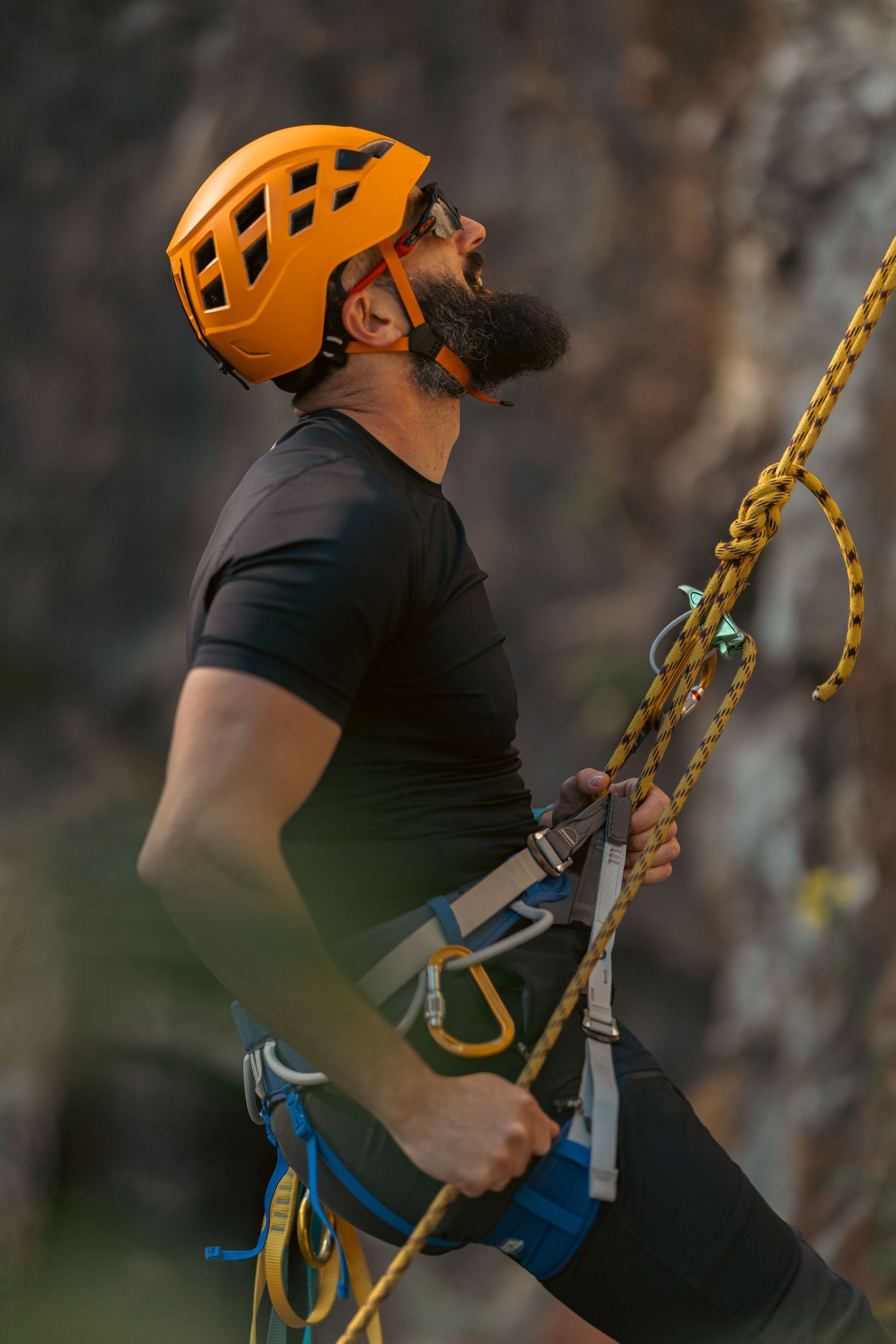 Bearded male rock climber in orange helmet and harness holding yellow rope, shouting with excitement outdoors.
