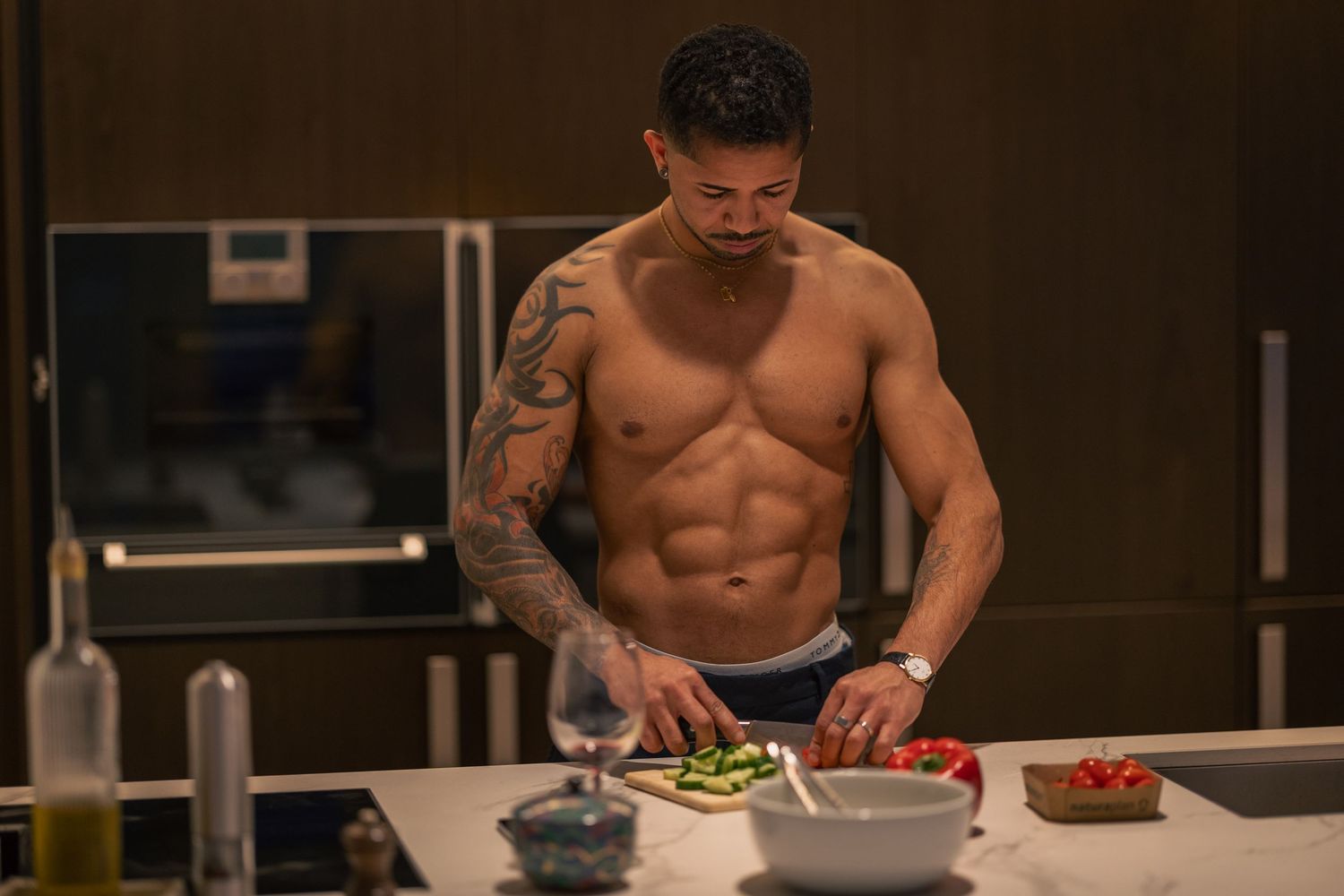 Shirtless muscular man with tattoos preparing fresh vegetables in a modern kitchen.
