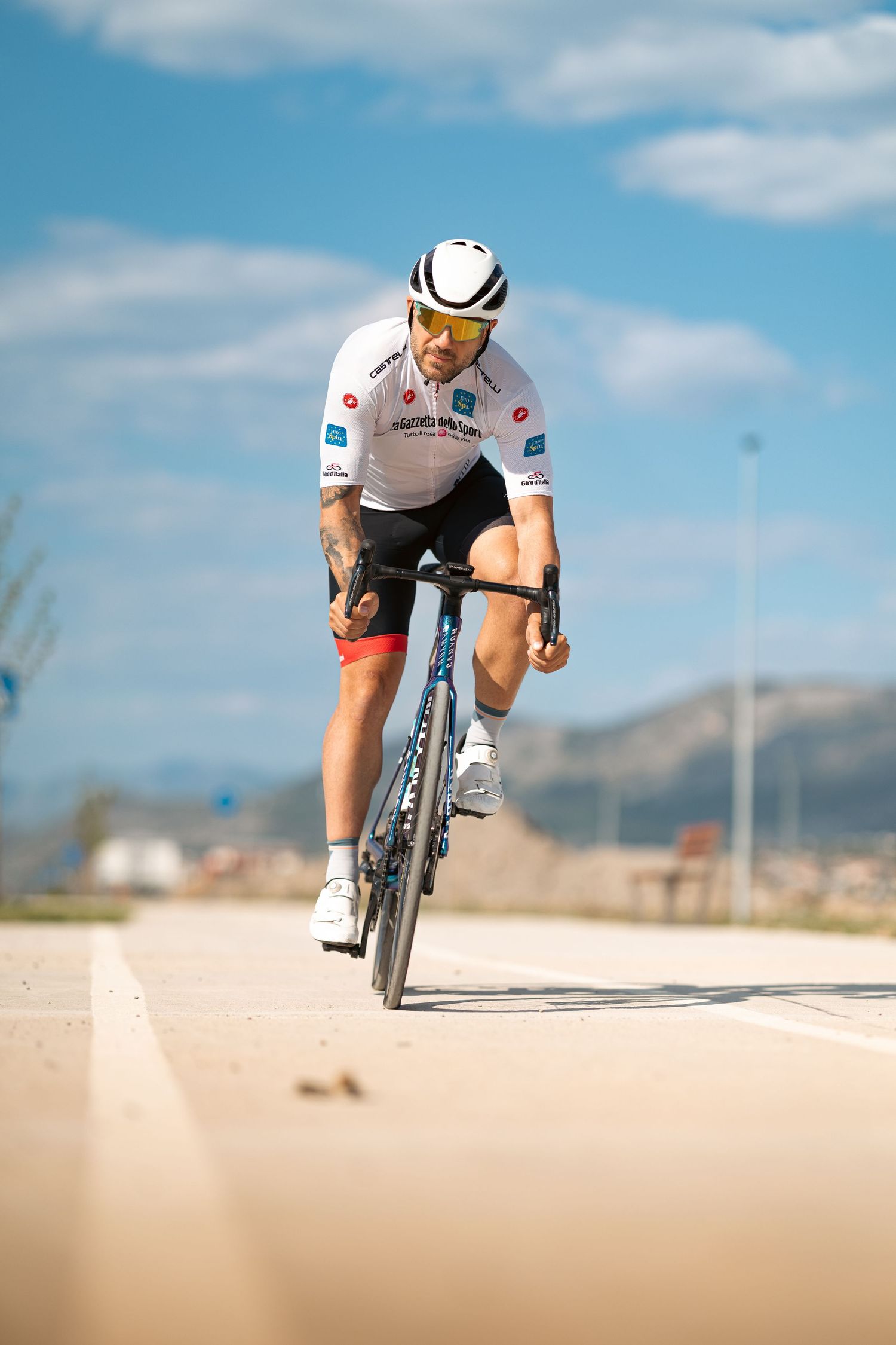 Competitive cyclist in white Astana jersey rides Canyon road bike on open road with mountains in background.