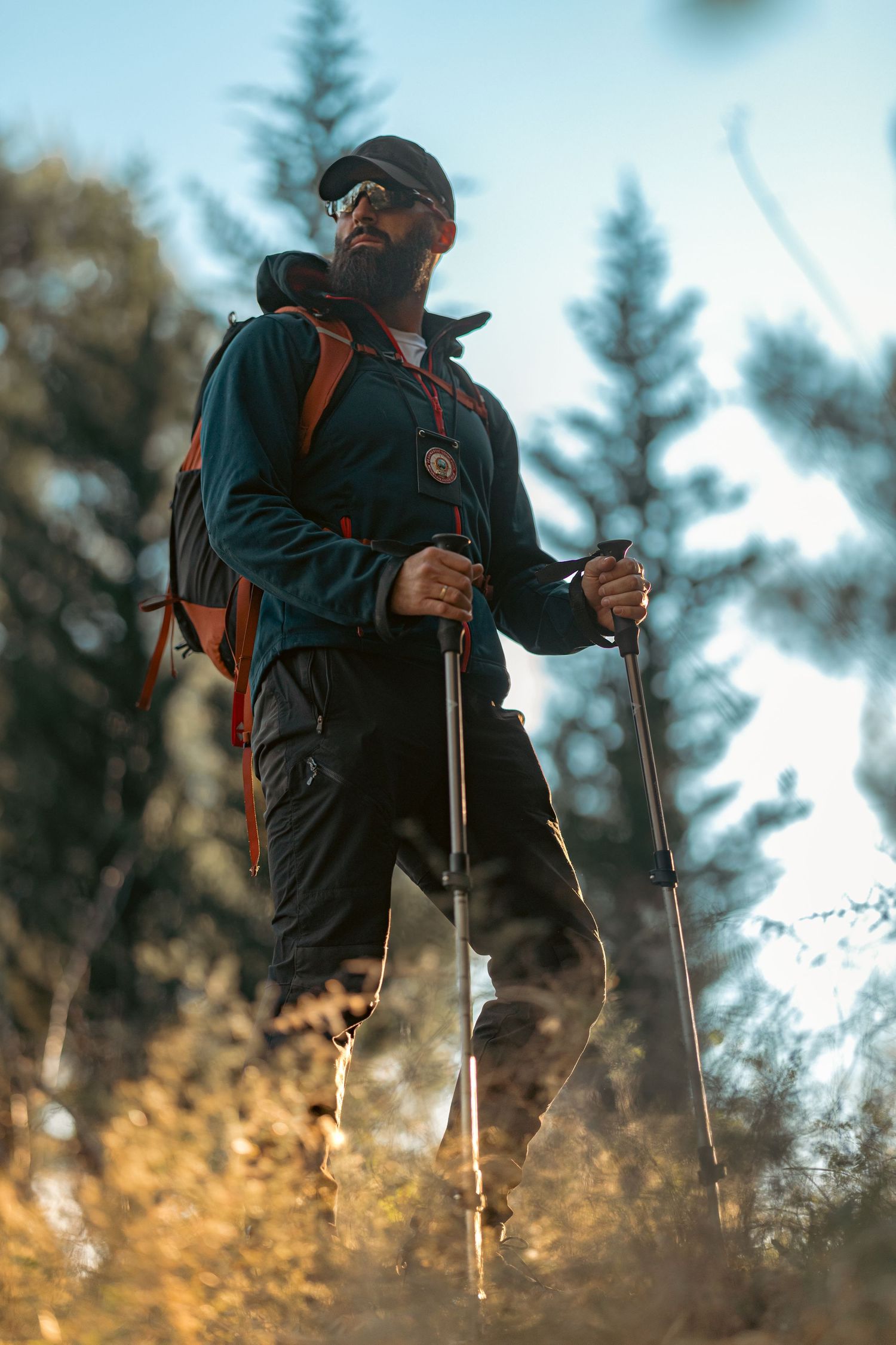 Bearded male hiker with trekking poles and orange backpack hiking through a forest trail on a sunny day.