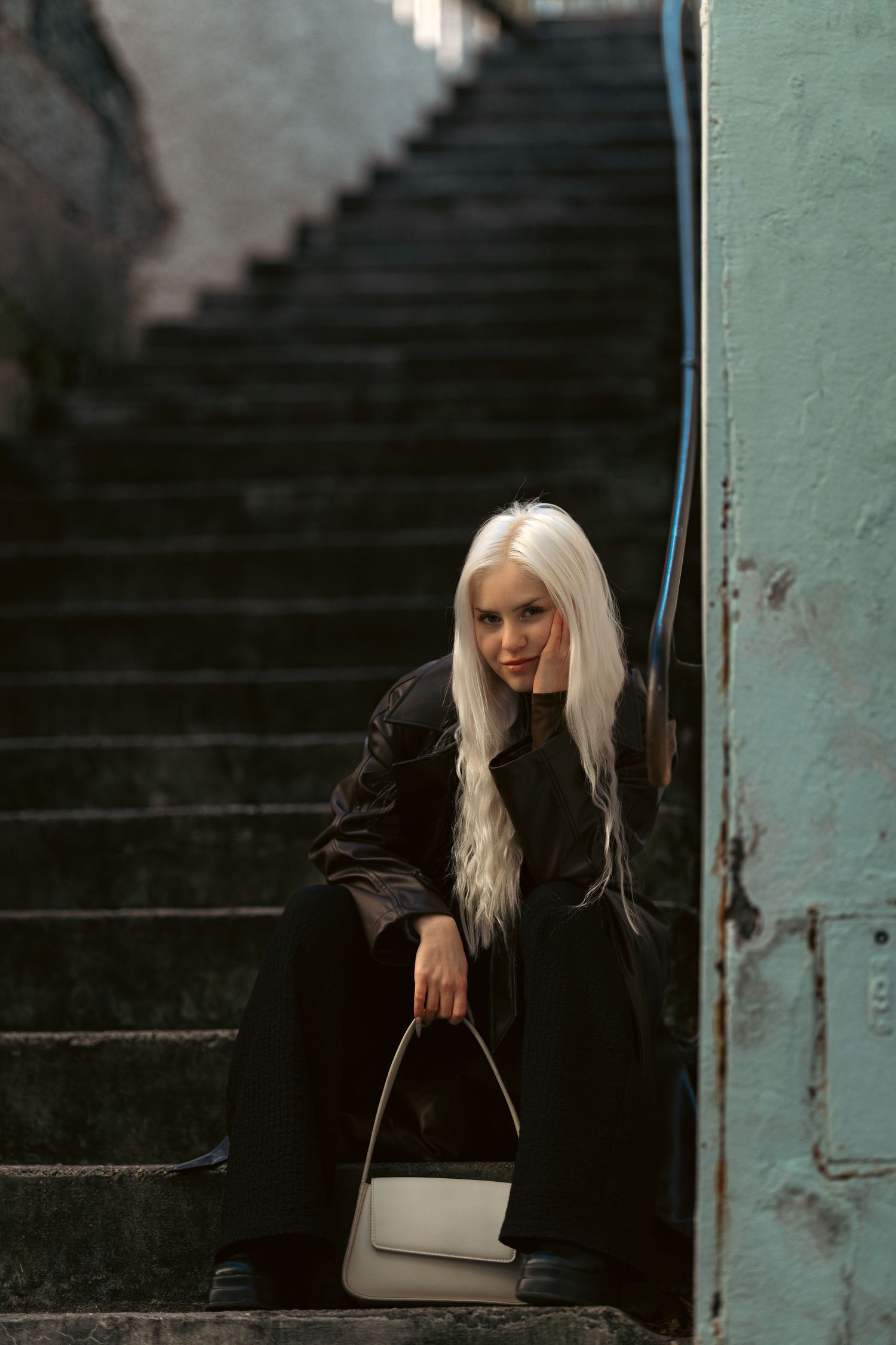 Young woman with long blonde hair in black outfit sits on dark stone stairs holding a white handbag.