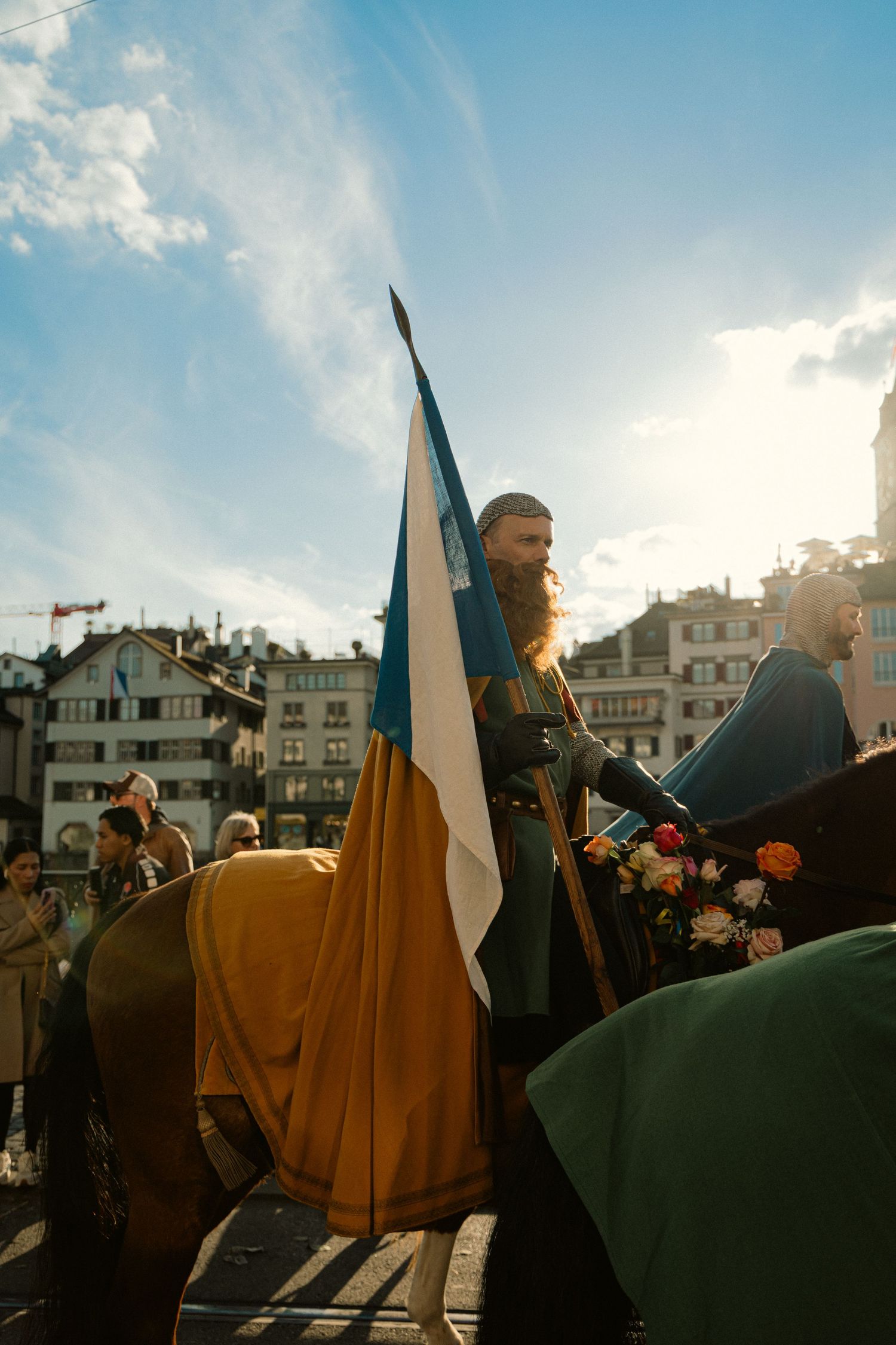Bearded man in medieval costume holding a blue and white flag on horseback at a sunny outdoor historical reenactment.