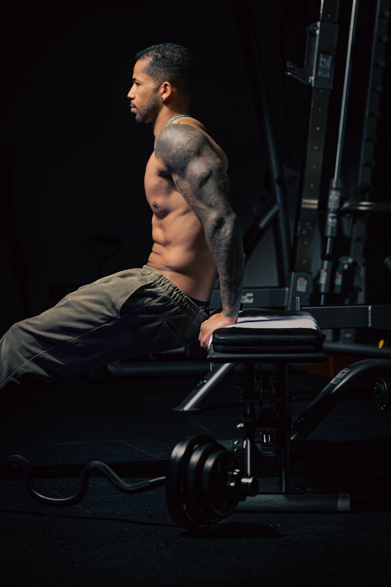 Muscular shirtless man with tattoos resting on gym bench beside barbell weights in dark gym.