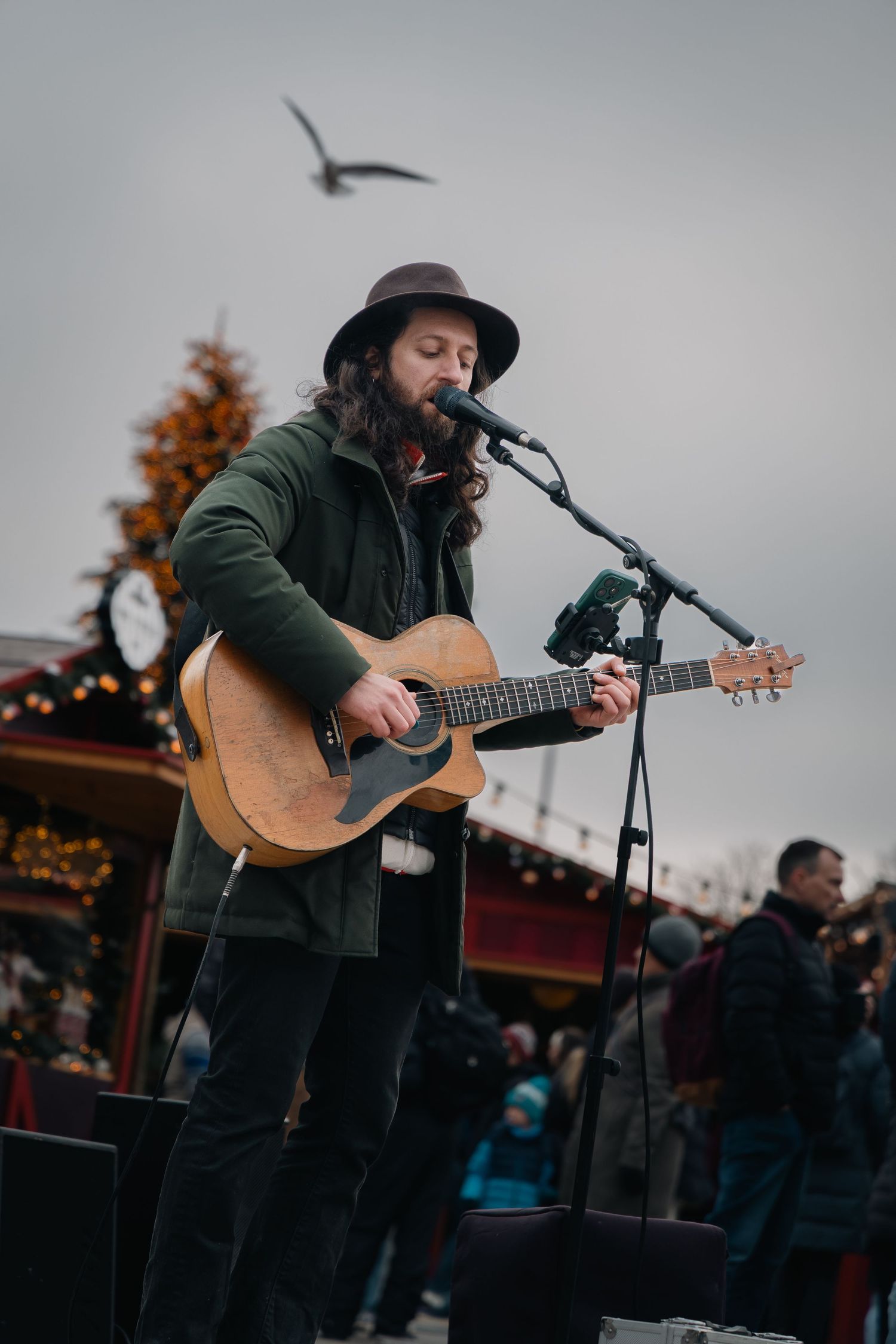 Musician performing with acoustic guitar outdoors at dusk with festive lights in background.