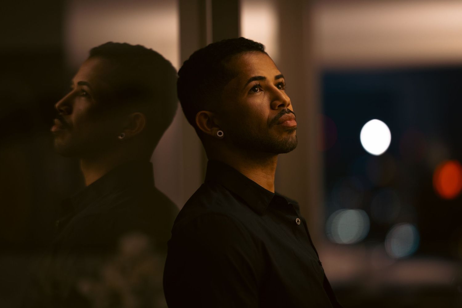 Young man in black shirt leaning against mirror in moody dark room with city lights bokeh background.