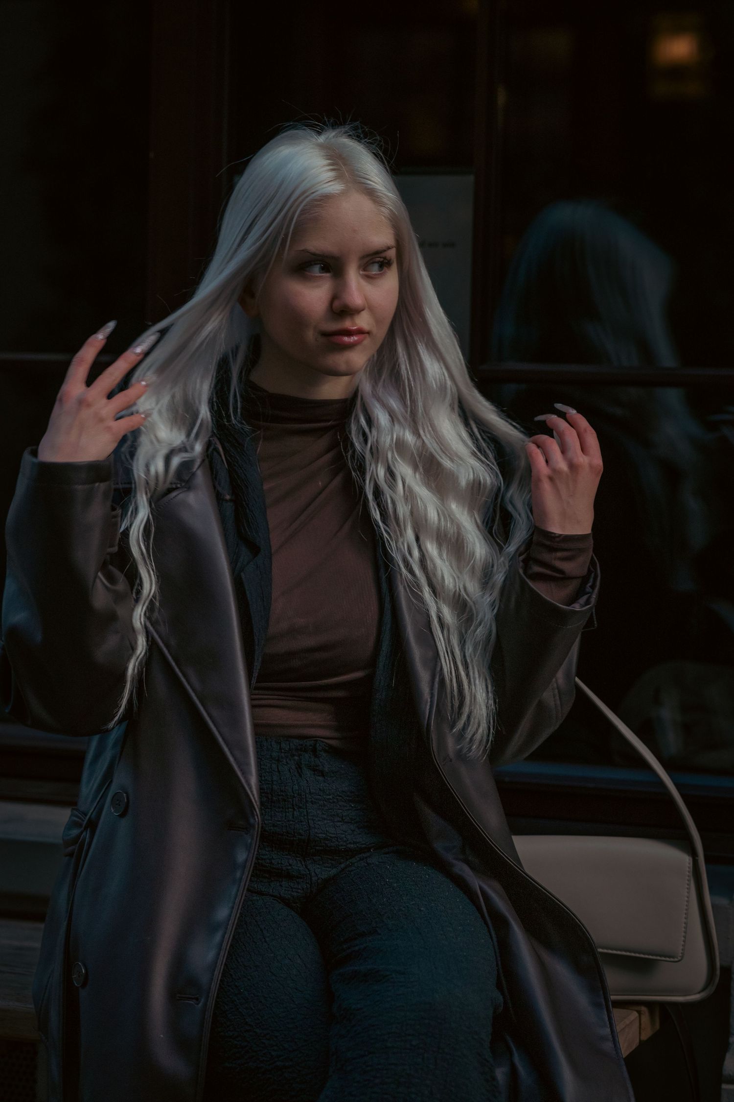 Person with long silver hair poses in leather jacket and brown top against dark background.