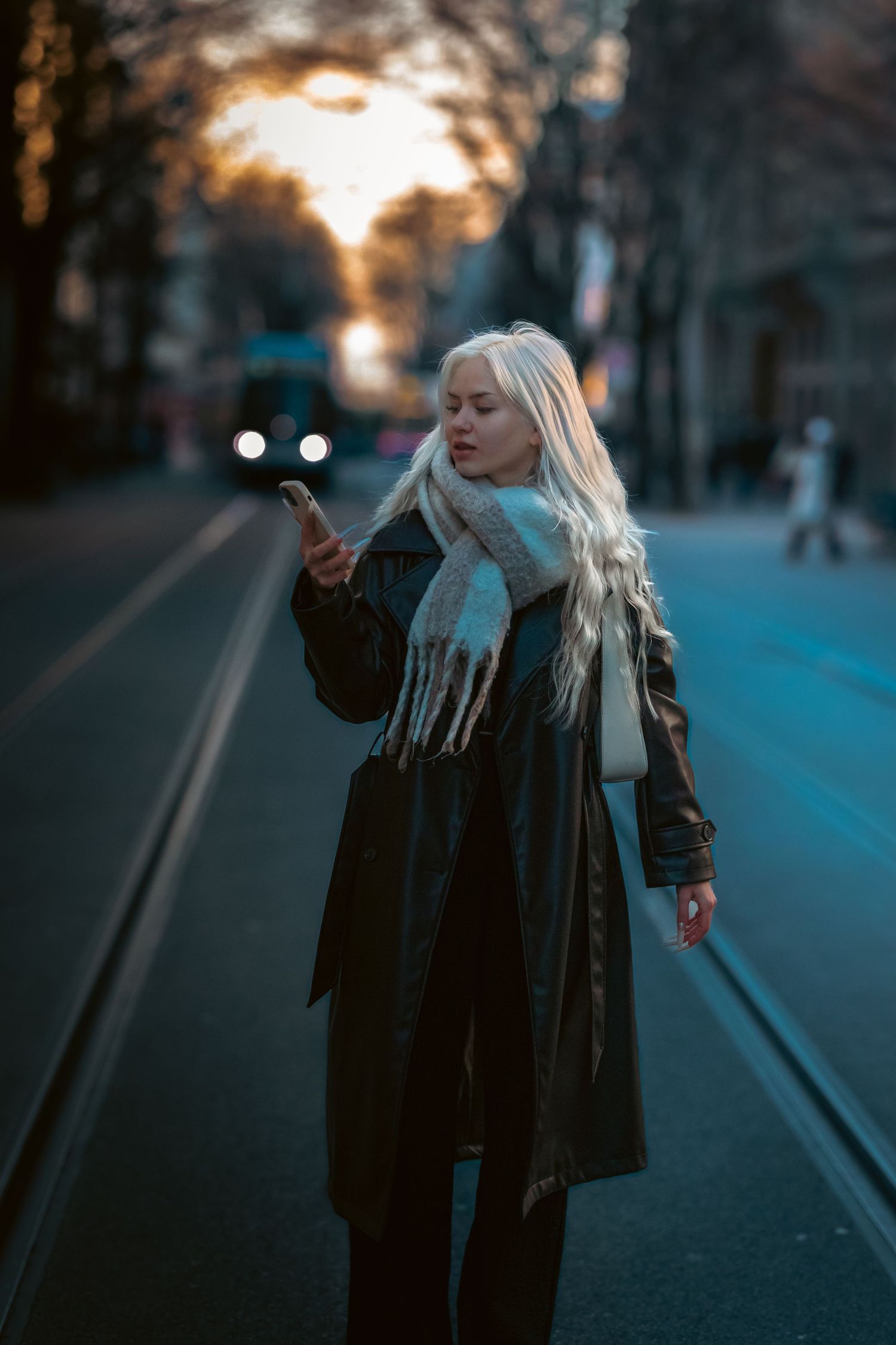 A figure with long white hair walks along a street at dusk wearing a black leather jacket.