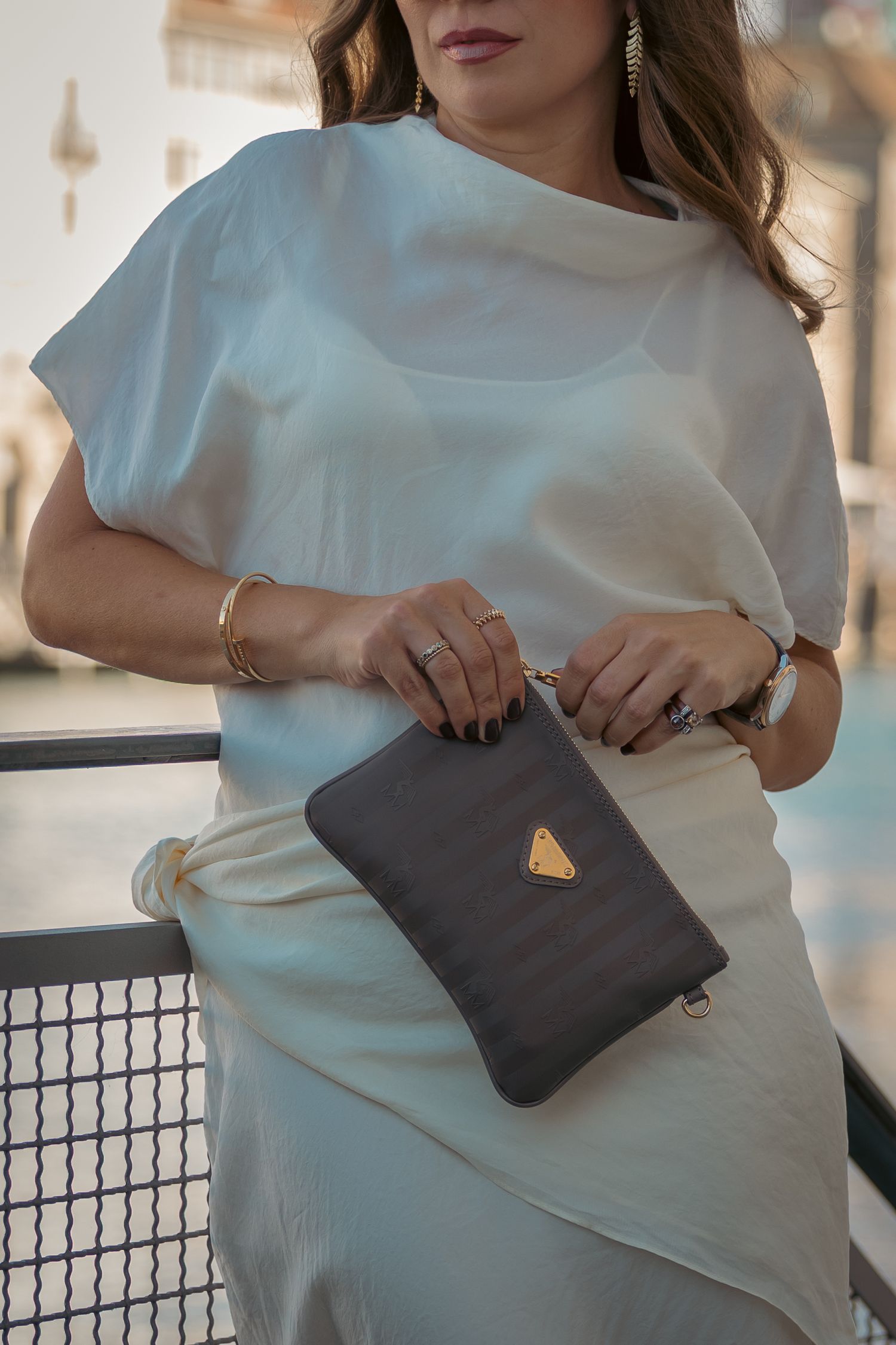 Woman in white outfit holding a dark gray clutch bag with gold triangle logo accent.
