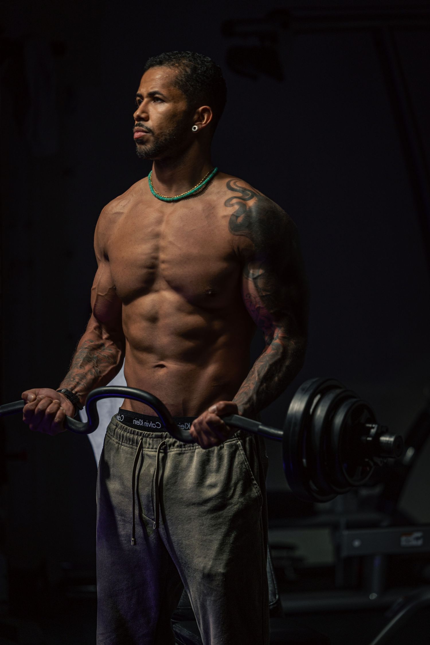 Muscular shirtless man with tattoos holds a barbell in a dark gym, wearing gray sweatpants and a green necklace.