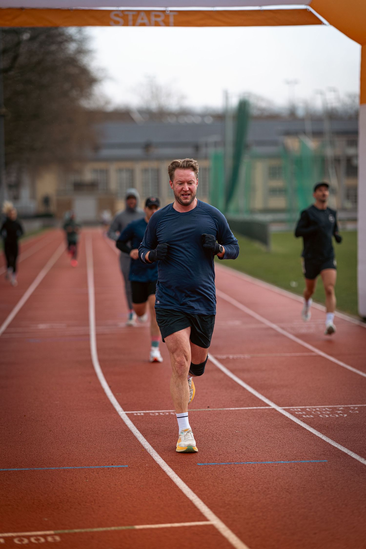 Runner sprinting on red running track during training session with others visible in background.