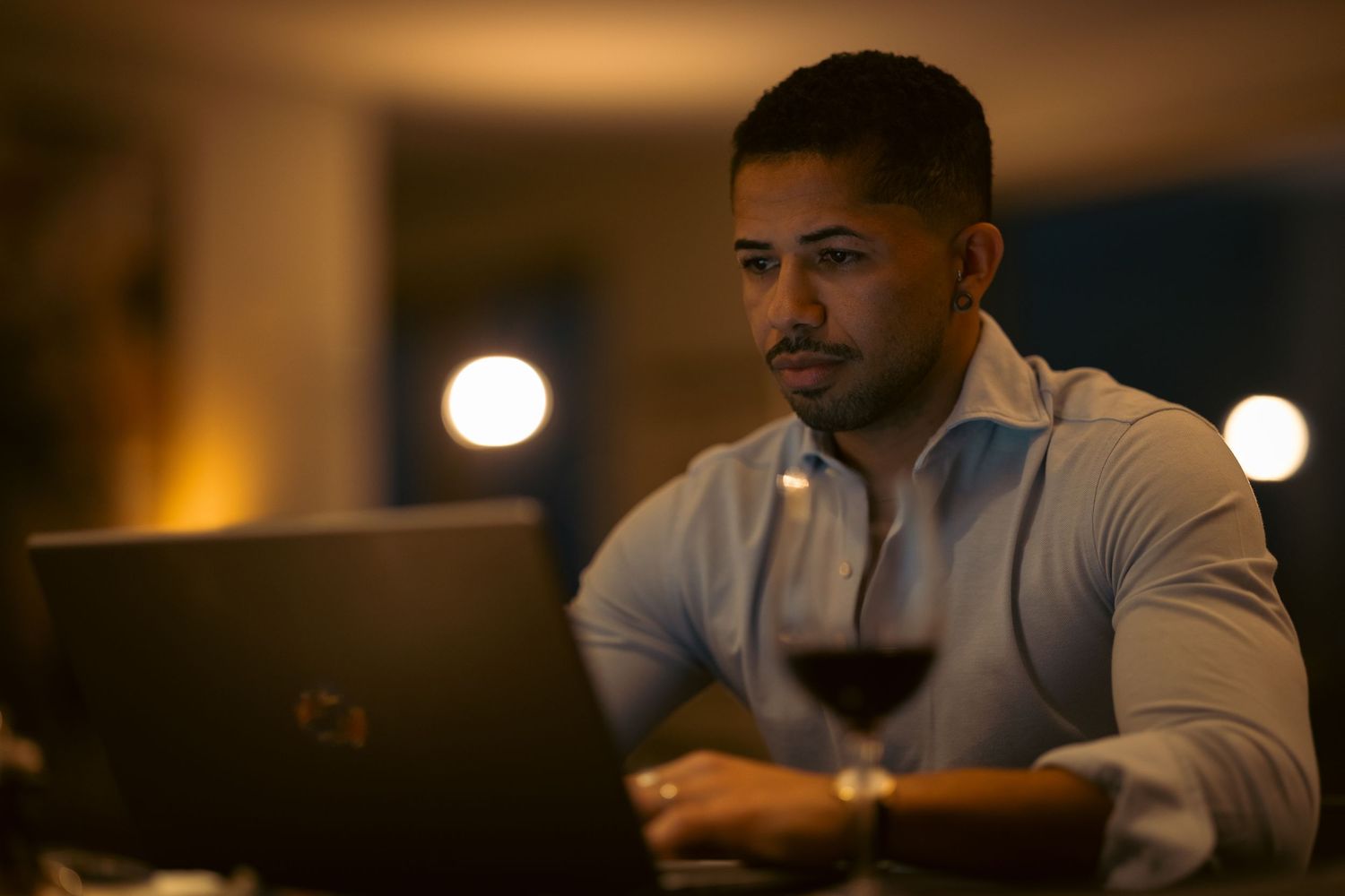 Man working on laptop at night with a glass of red wine, focused expression in warm ambient lighting.