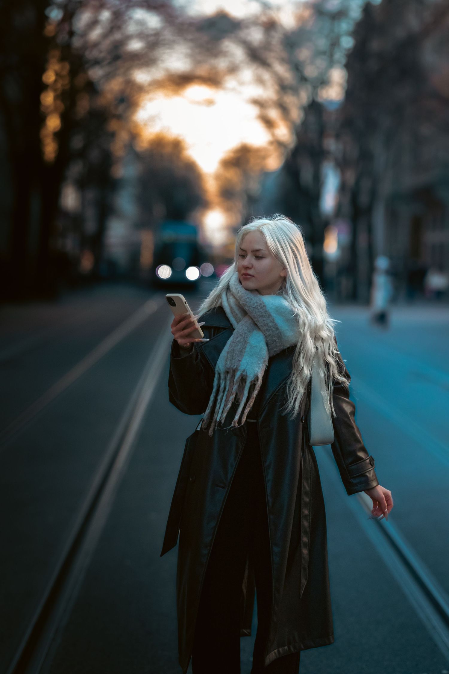 Person in winter coat and scarf checks phone on empty street at dusk with blurred bokeh lights in background.
