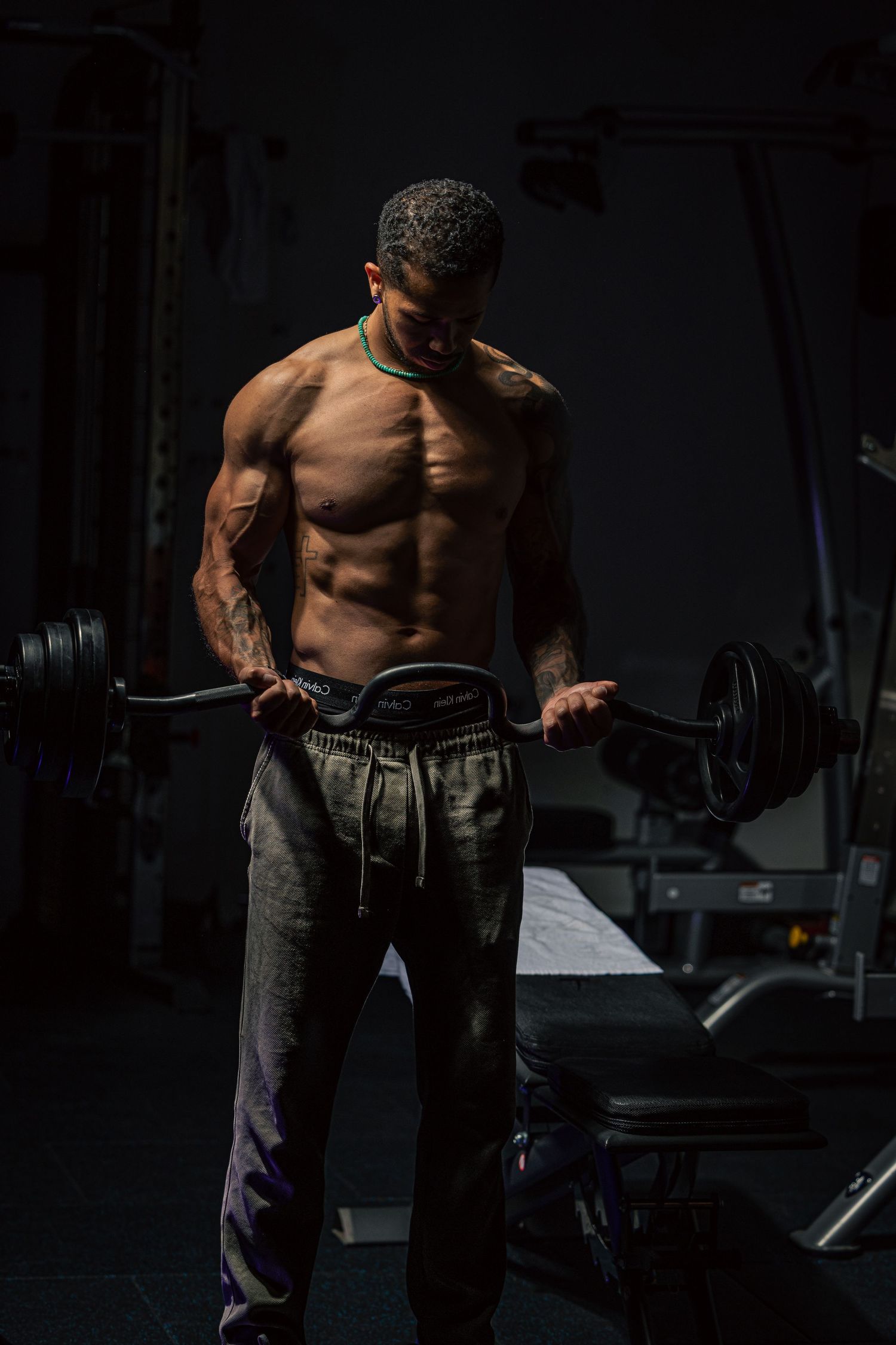 Muscular shirtless man with tattoos holding a barbell in a dark gym, showcasing defined abs and physique.