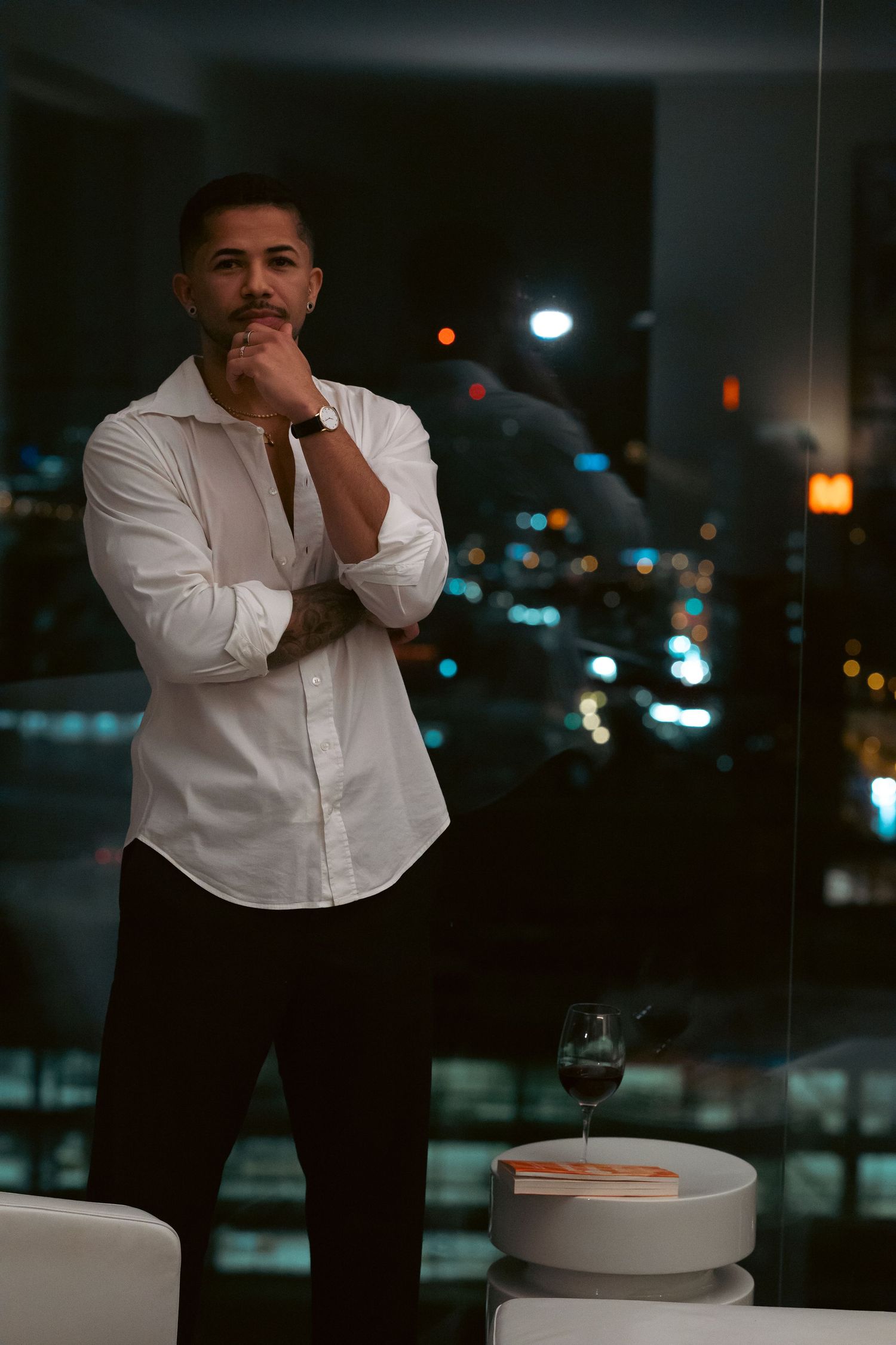 A confident man in a white shirt stands thoughtfully by a window with a city night skyline backdrop.