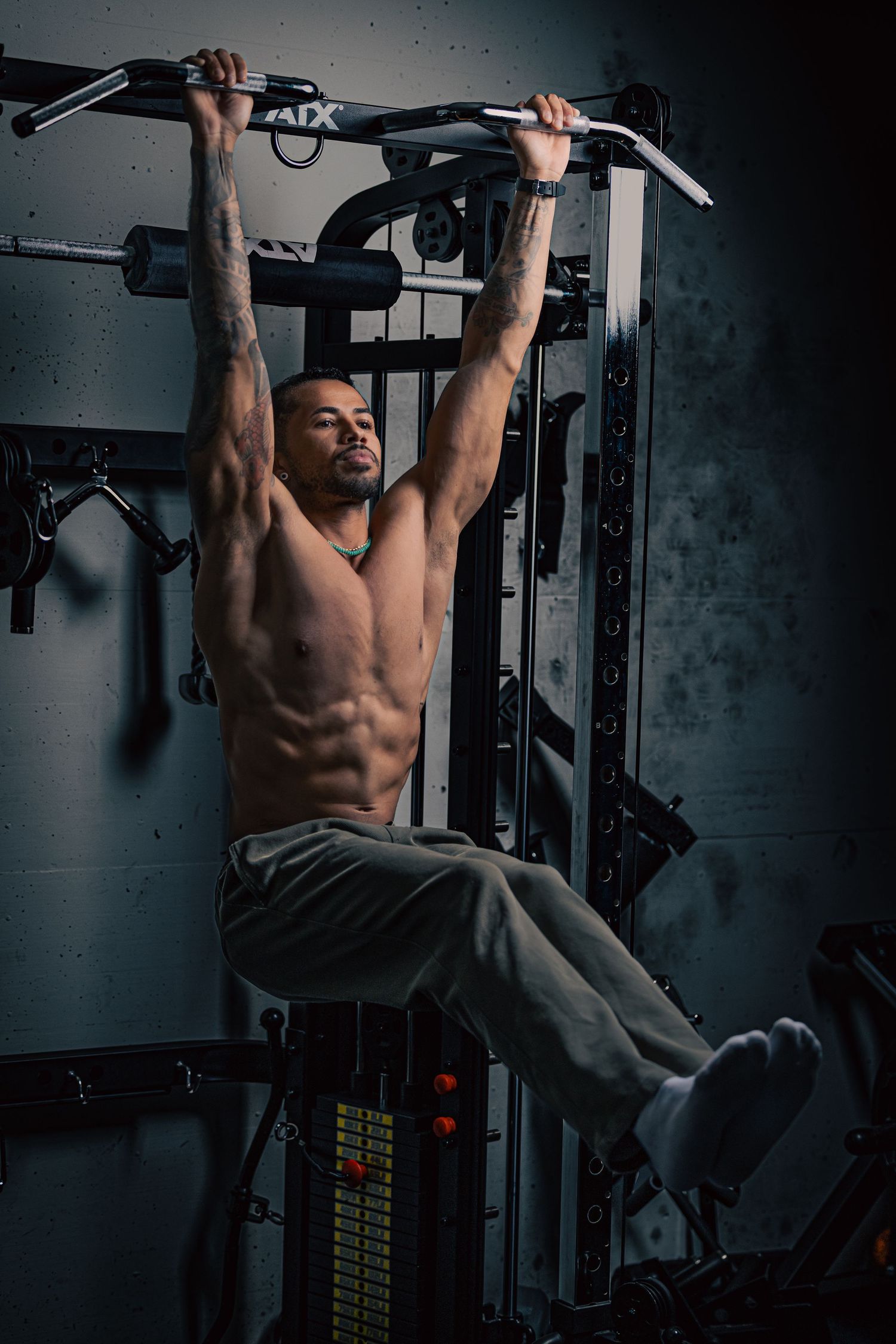 Shirtless muscular man doing pull-ups on an ATX power rack, displaying defined abs and tattooed arms in a dark gym.