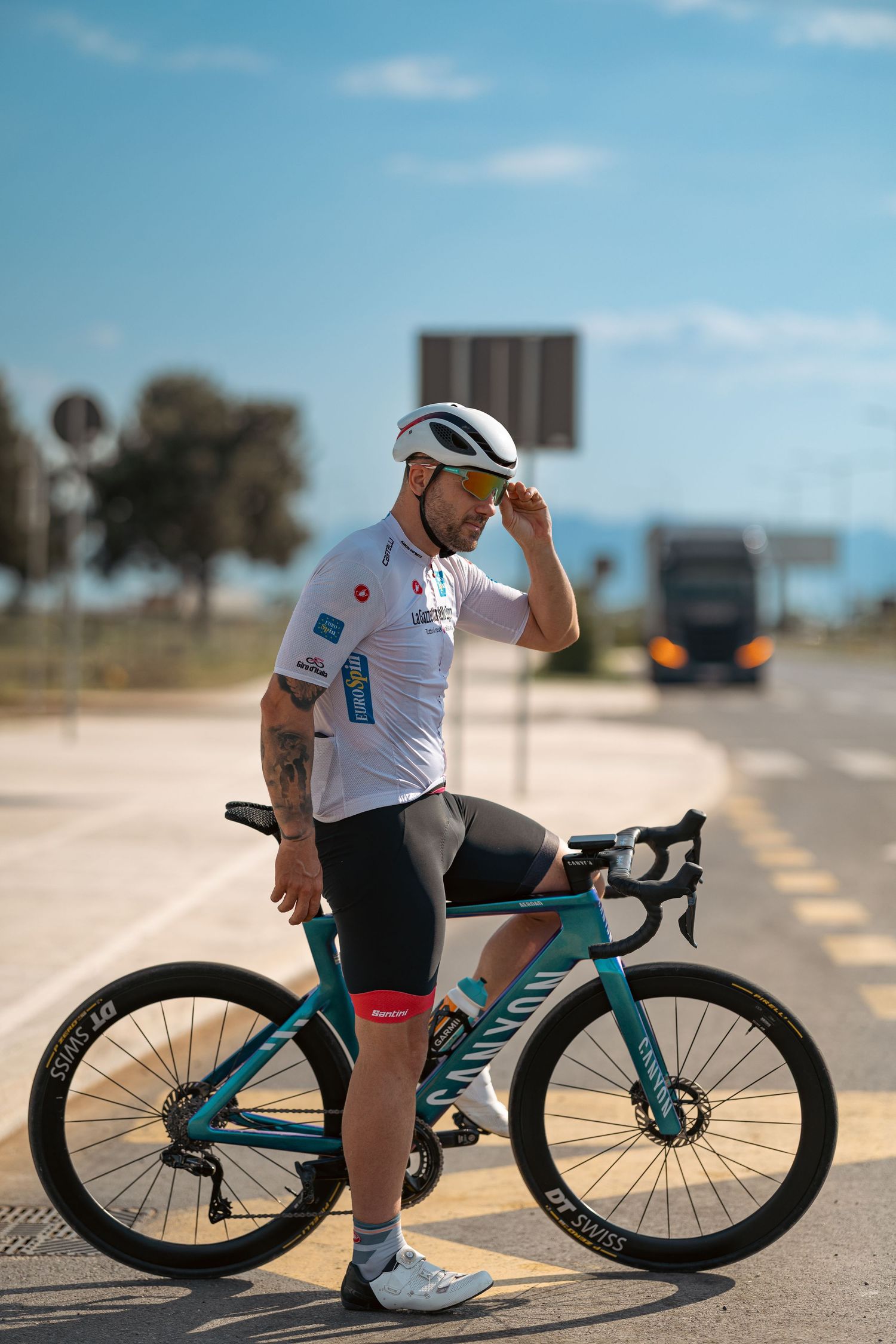 Professional cyclist in white jersey and helmet stands beside a teal road bike on a sunny day.