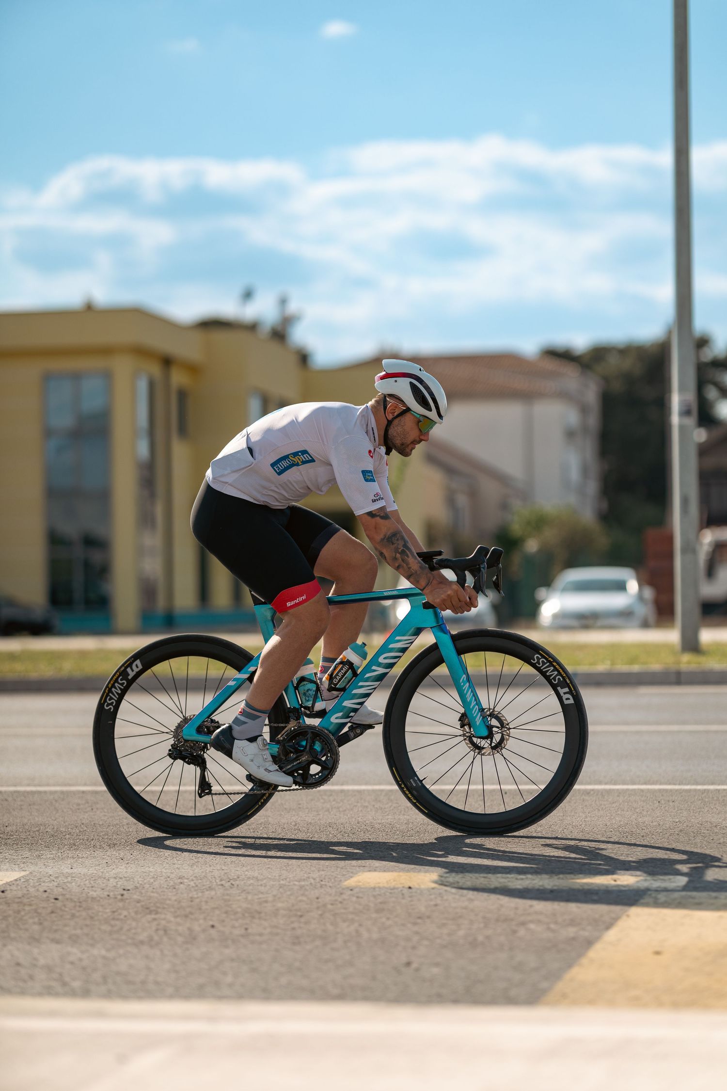 Cyclist in white team jersey rides teal Canyon bike through urban street intersection on a sunny day.