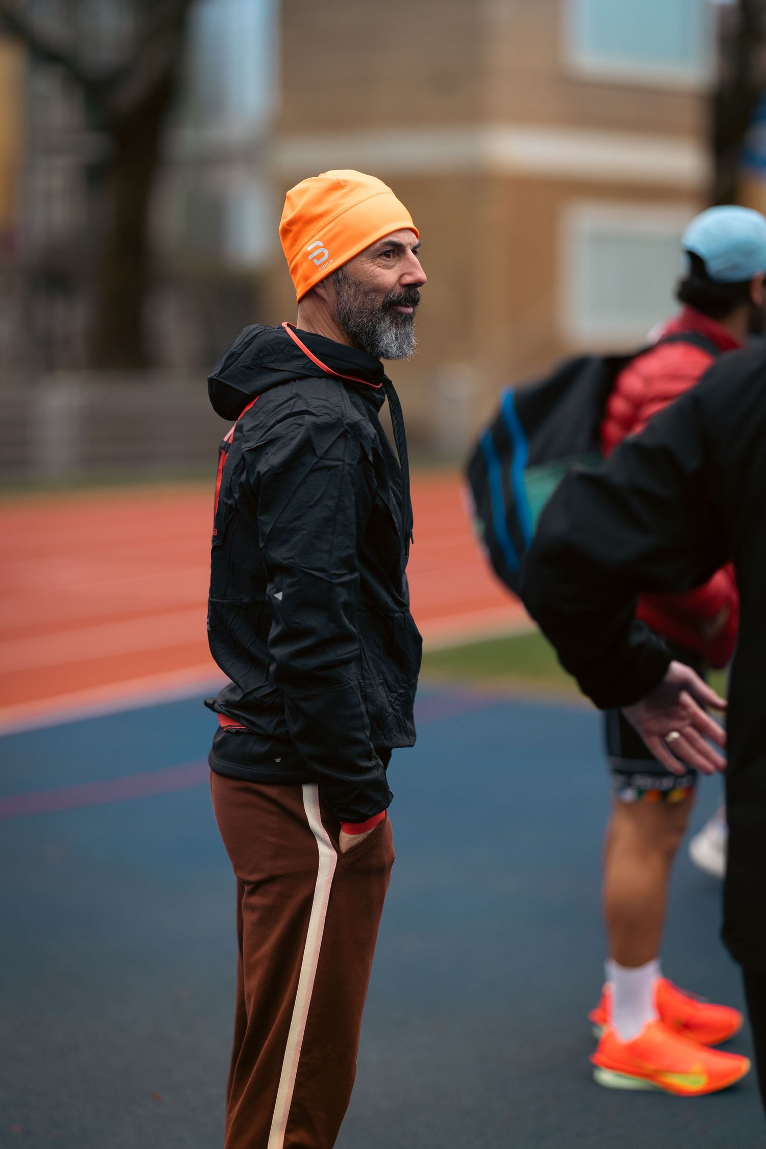 Runner wearing an orange beanie and black jacket standing on an outdoor track.