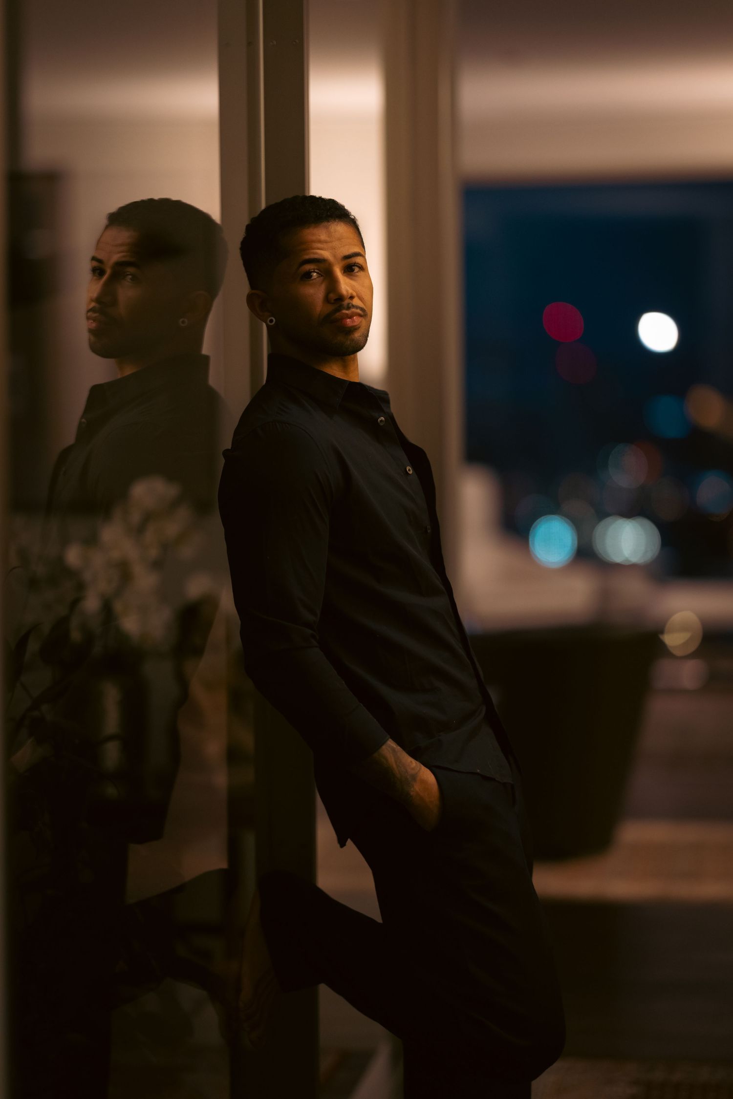 A well-dressed man in a black shirt leans against a glass wall at night, his reflection visible beside him.