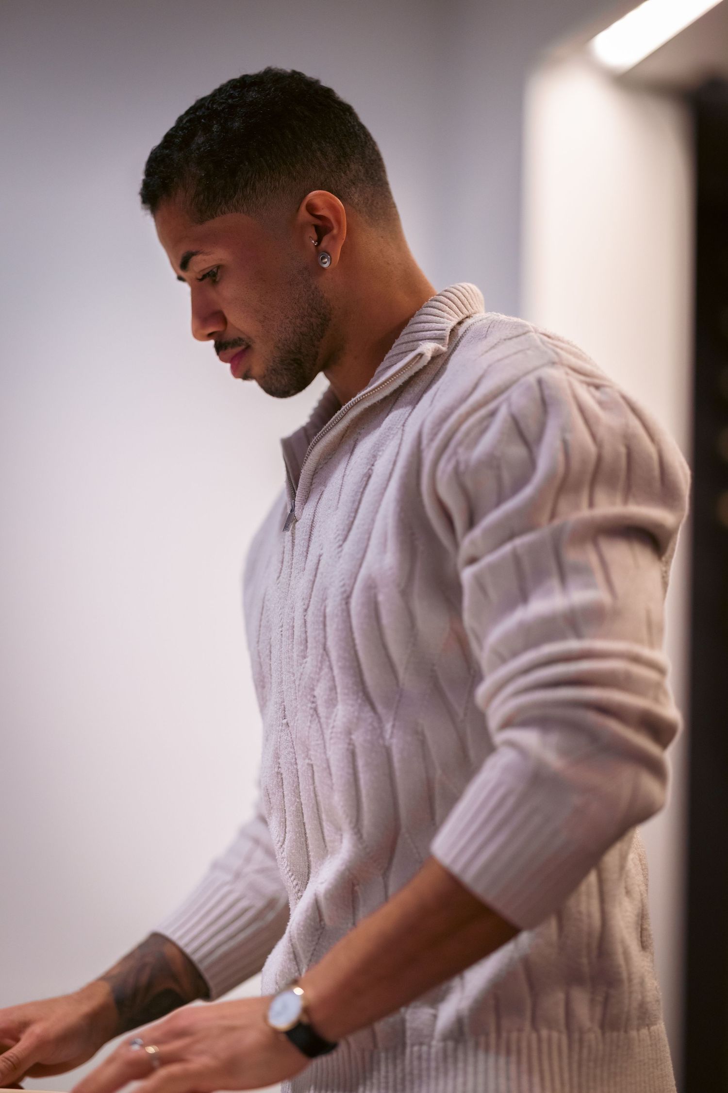 Young man in a cream cable-knit zip sweater with ear stud and watch, looking down thoughtfully.