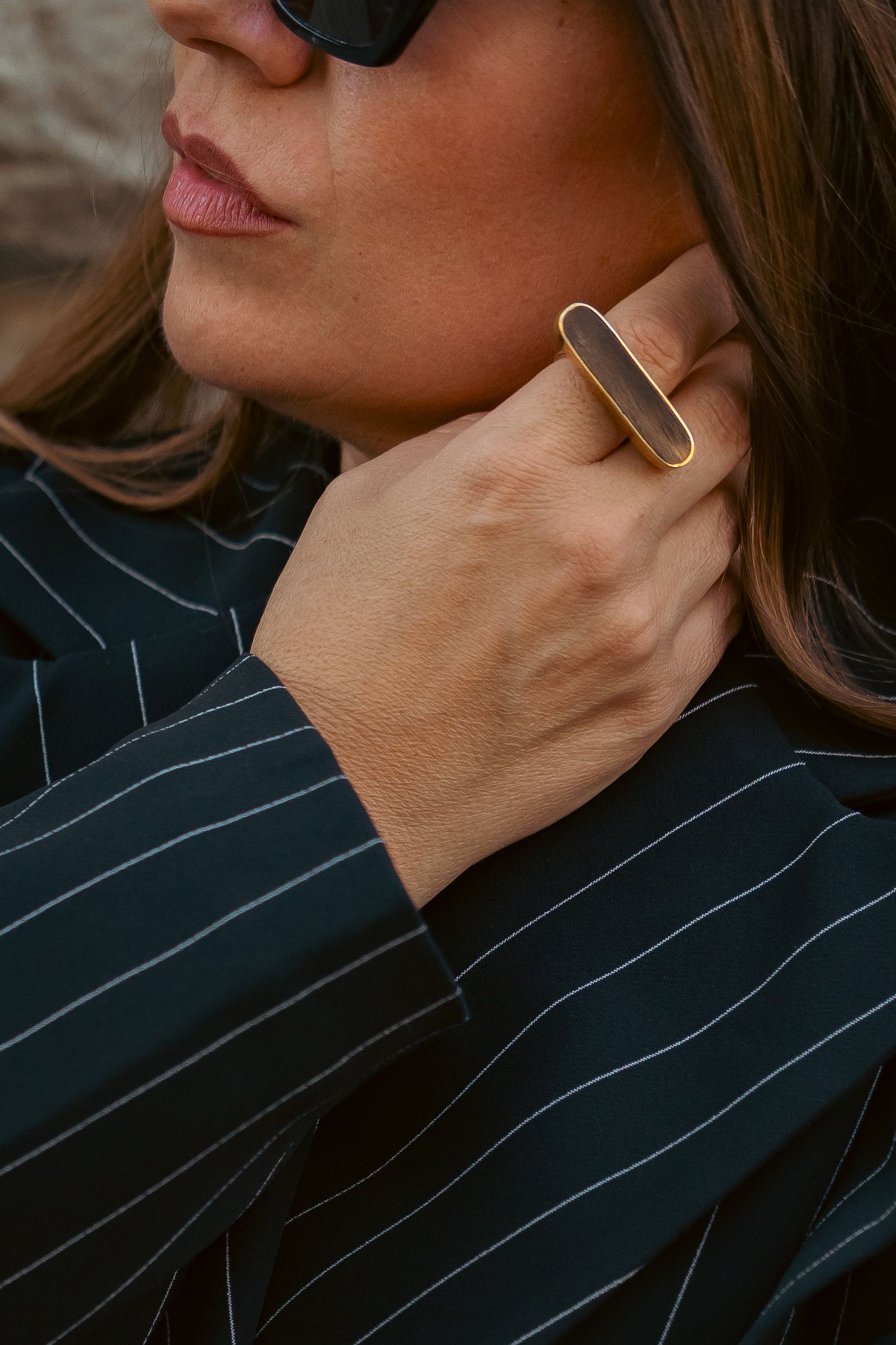 Woman wearing a gold rectangular ring with dark stone, paired with a navy pinstripe blazer and black sunglasses.