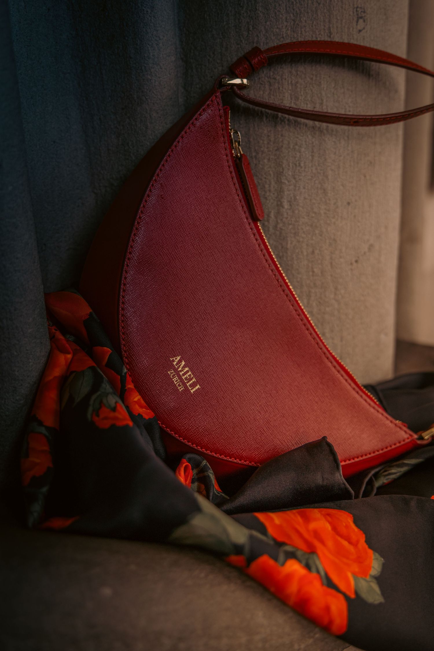 A deep red AMELI crescent-shaped leather handbag resting against a floral scarf on a gray chair.