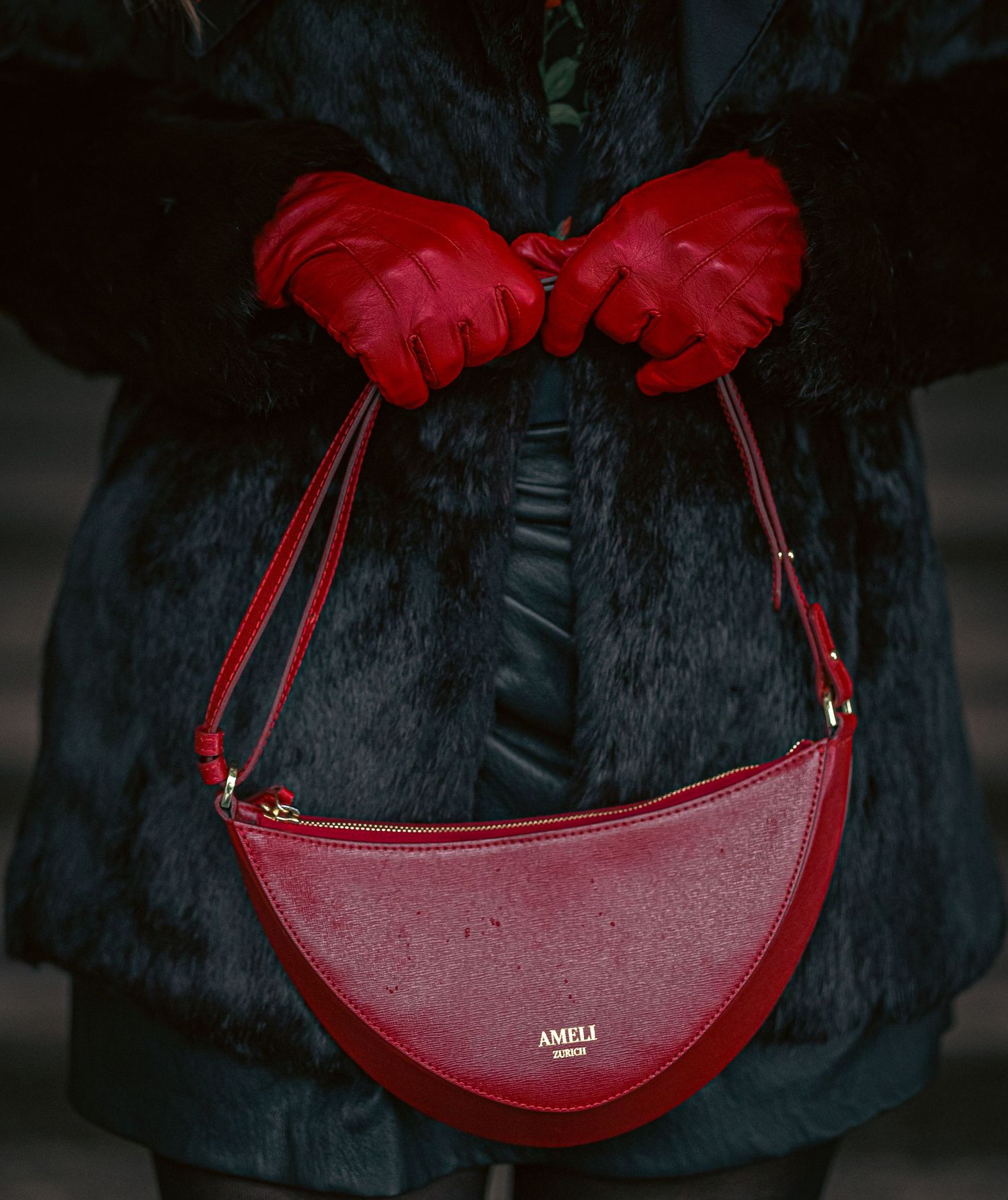 Red AMELI leather crescent shoulder bag held by a person wearing red gloves and a dark fur coat.