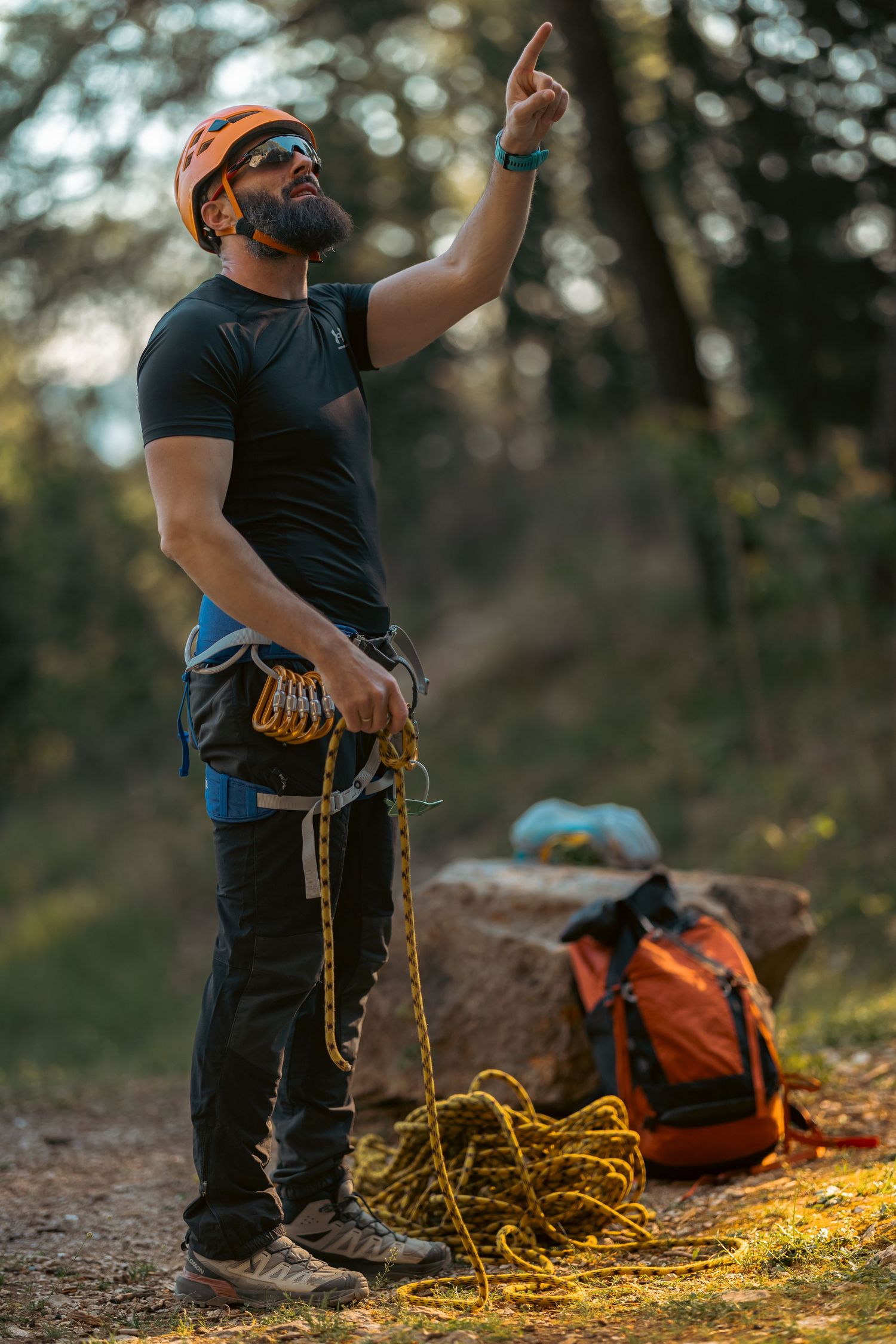 A bearded male rock climber in an orange helmet holds rope and points upward in a forest setting.