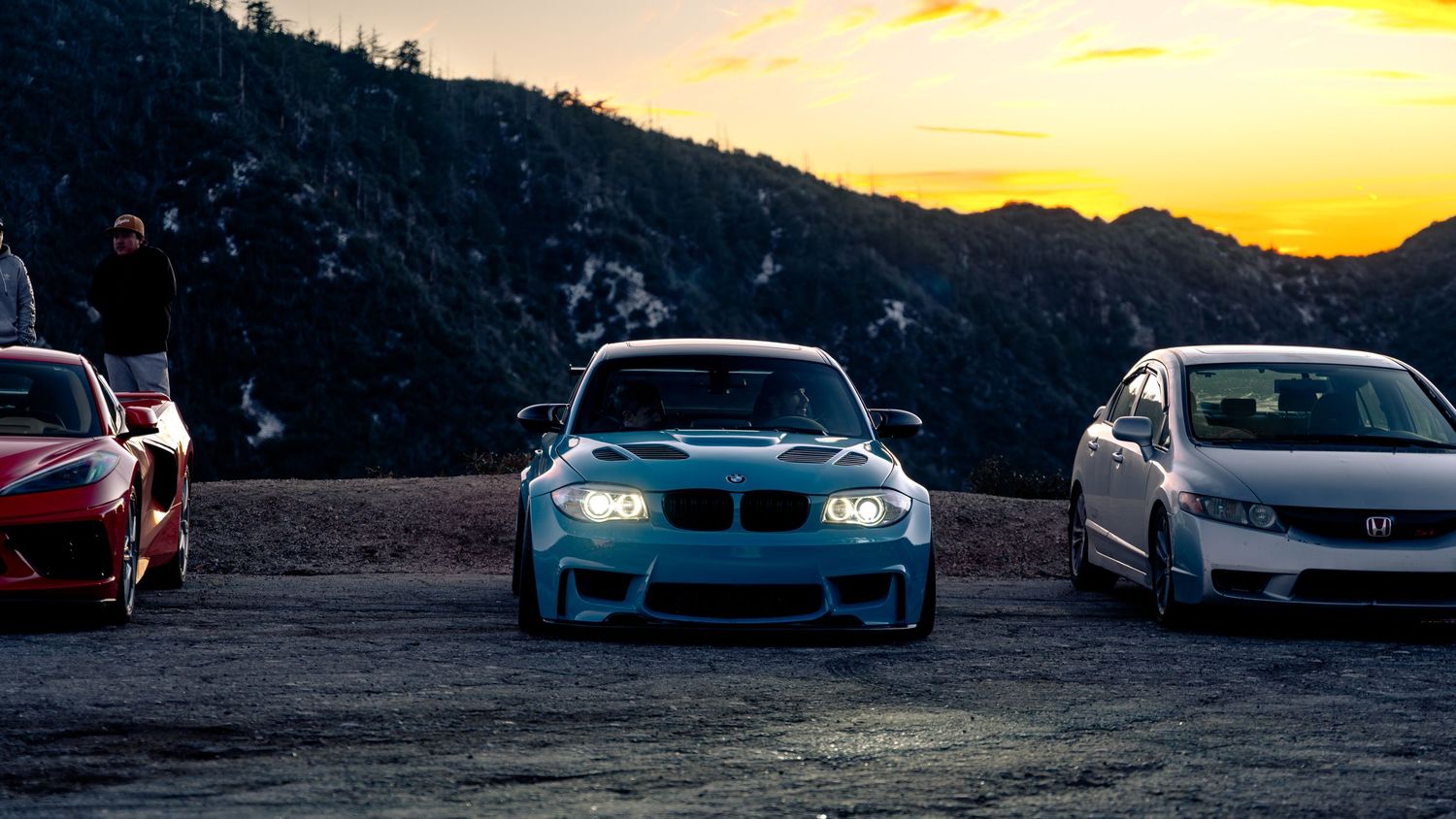 Exotic cars parked along mountain roadside during vibrant sunset.