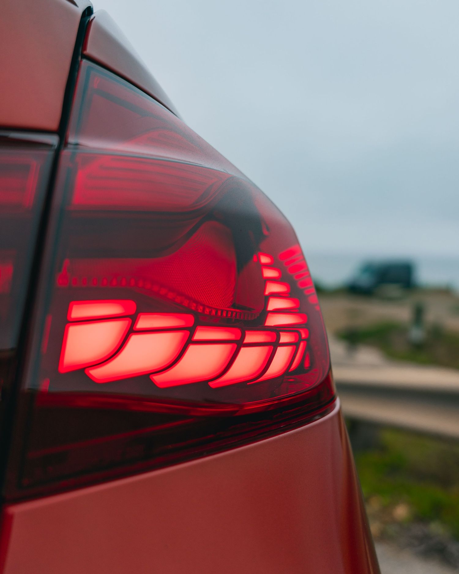Close-up of a red car's glowing LED tail light with layered design, parked near a coastal road.