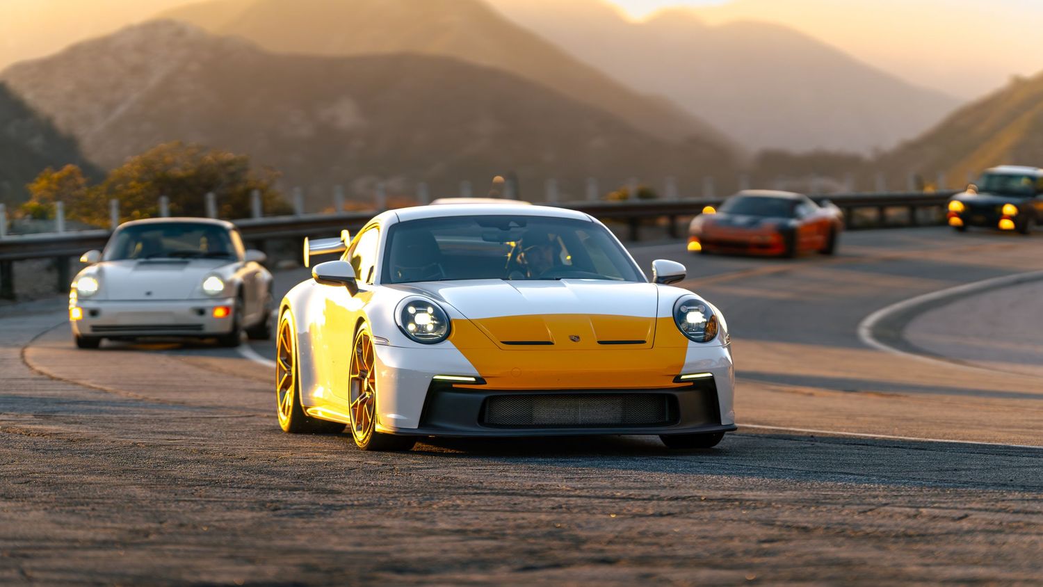 Row of luxury sports cars parked against mountain landscape at sunset.