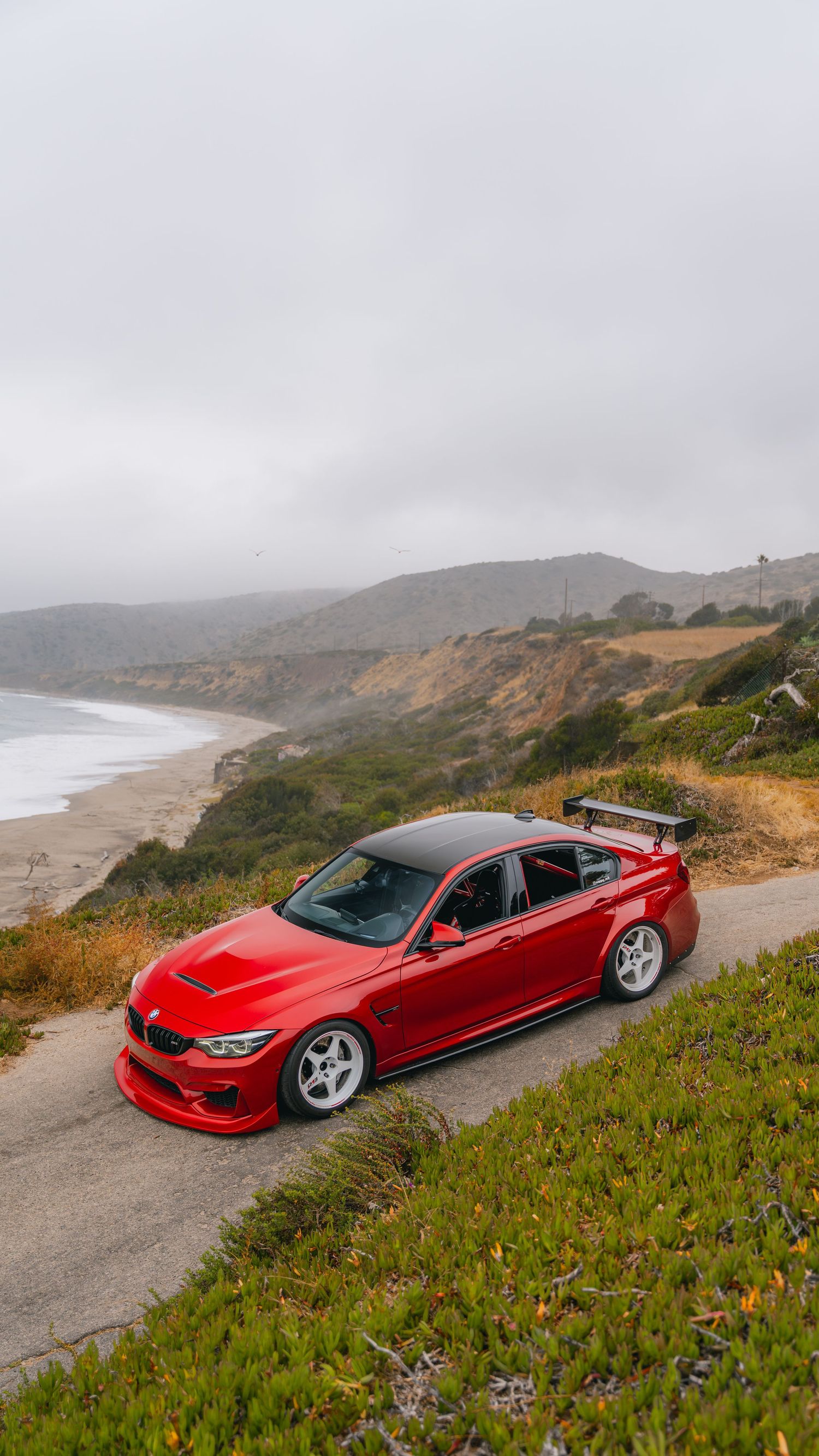 Red BMW M3 with large rear spoiler parked on coastal road overlooking misty beach and hills.